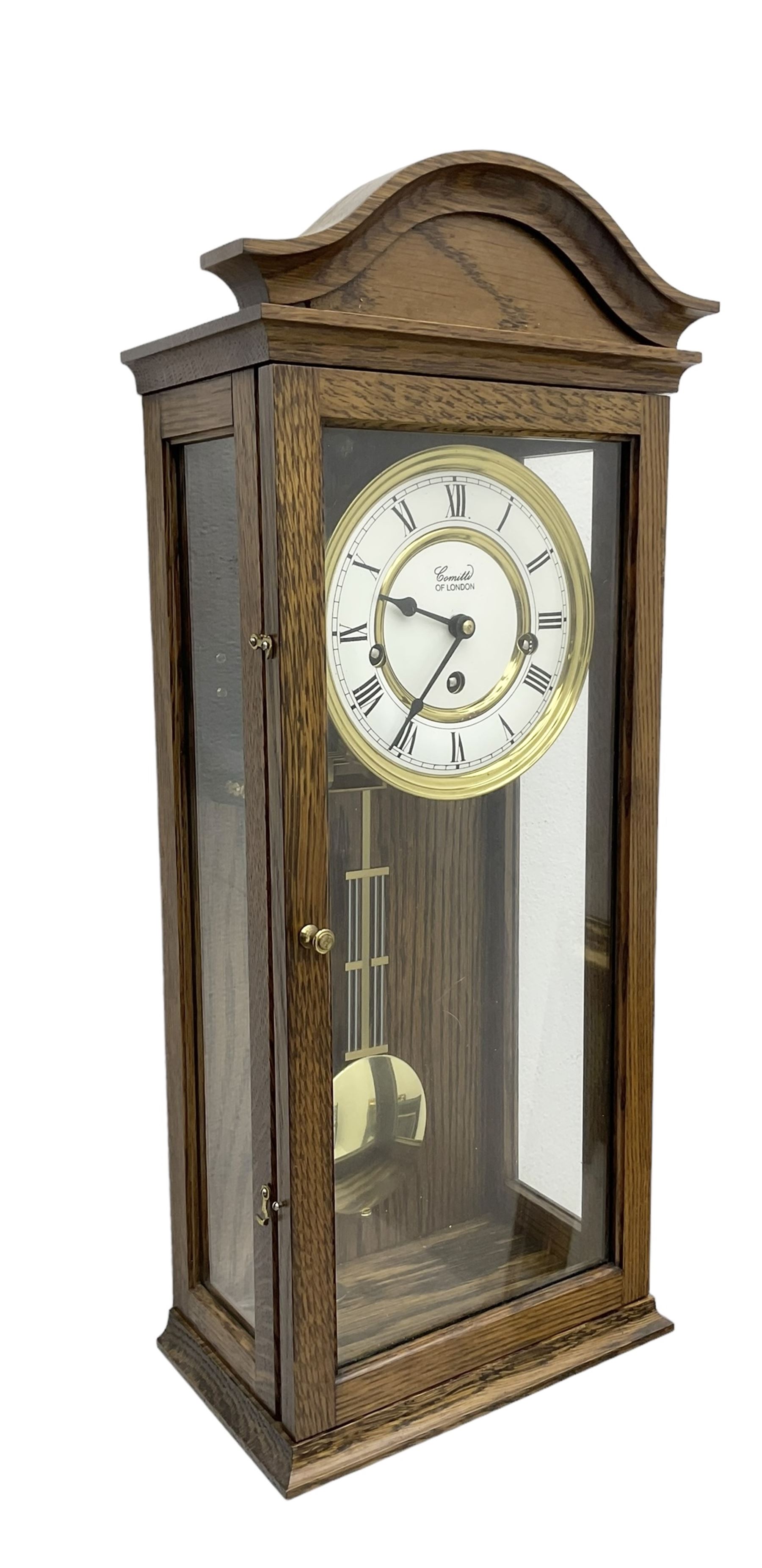 Commiti of London -  three train spring driven 8-day wall clock in an oak case, with a fully glazed door and curved pediment, two part dial with Roman numerals, spade hands and gridiron pendulum, rack movement chiming the hours and quarters on 5 gong rods. With key.