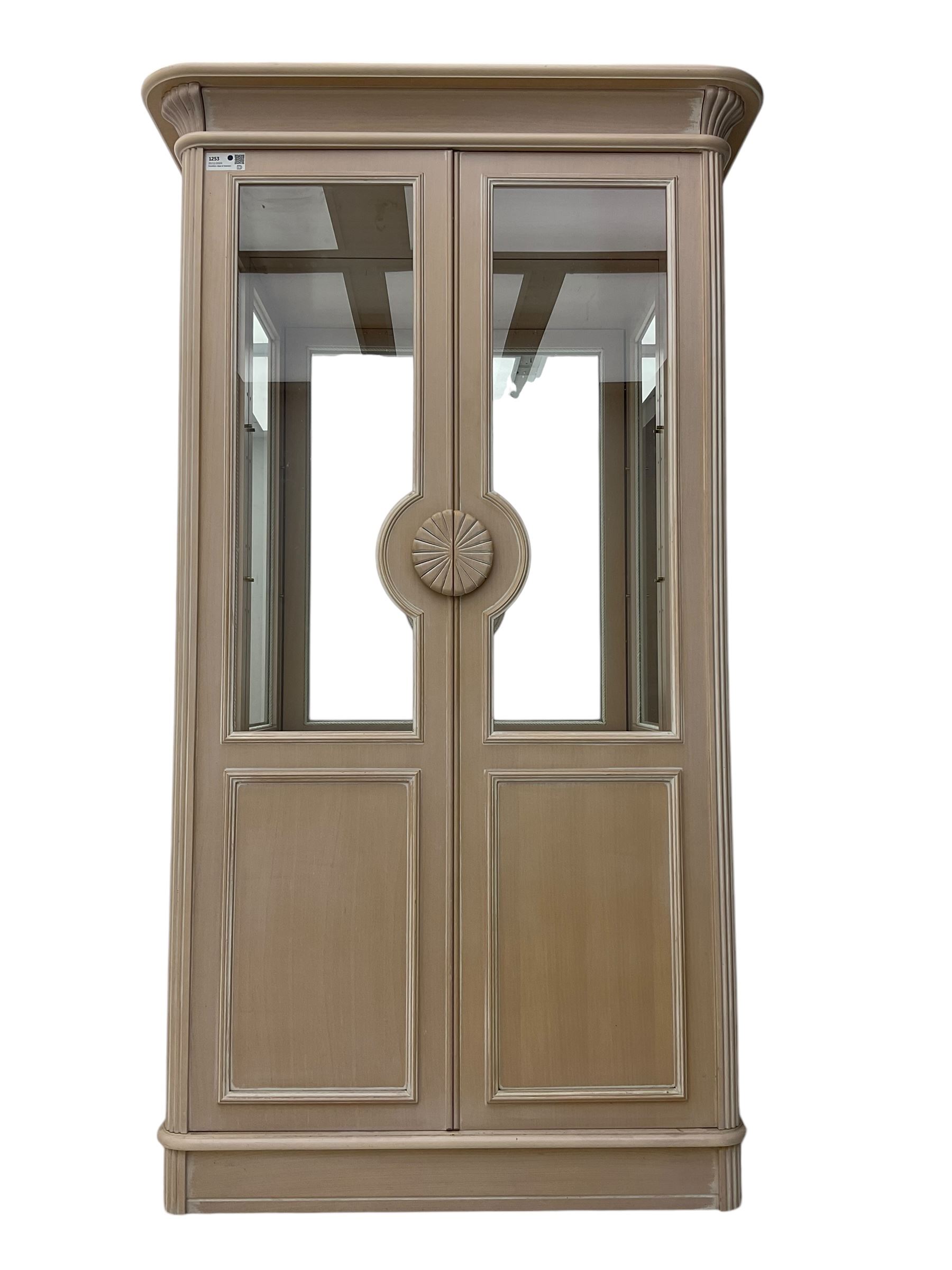 Contemporary display cabinet, moulded cornice above glazed panelled doors with central carved sunburst handles, mirrored back interior with lighting, lower enclosed storage compartment, raised on plinth base