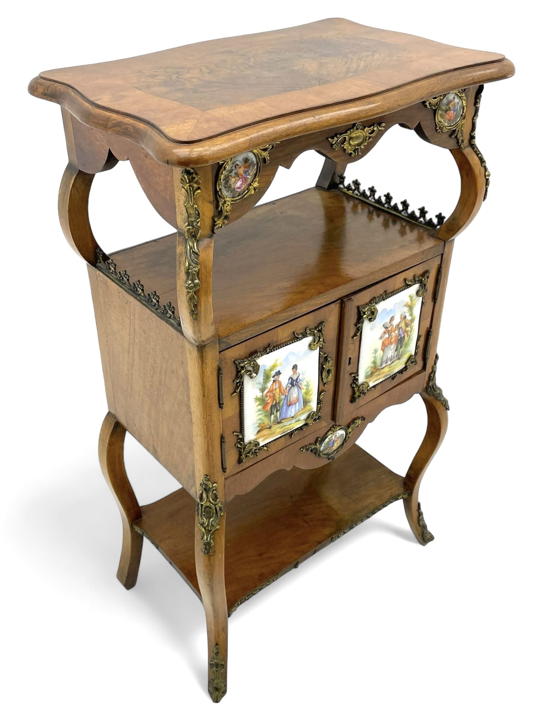 Mid-to-late 20th century French figured walnut three-tier bedside stand, shaped moulded top inlaid with figured book-matched veneers and crossbanding, double cupboard below enclosed by two doors mounted by painted porcelain panels depicting courting scenes within landscapes, on cabriole supports united by undertier, decorated with ornate cast metal cartouche mounts 