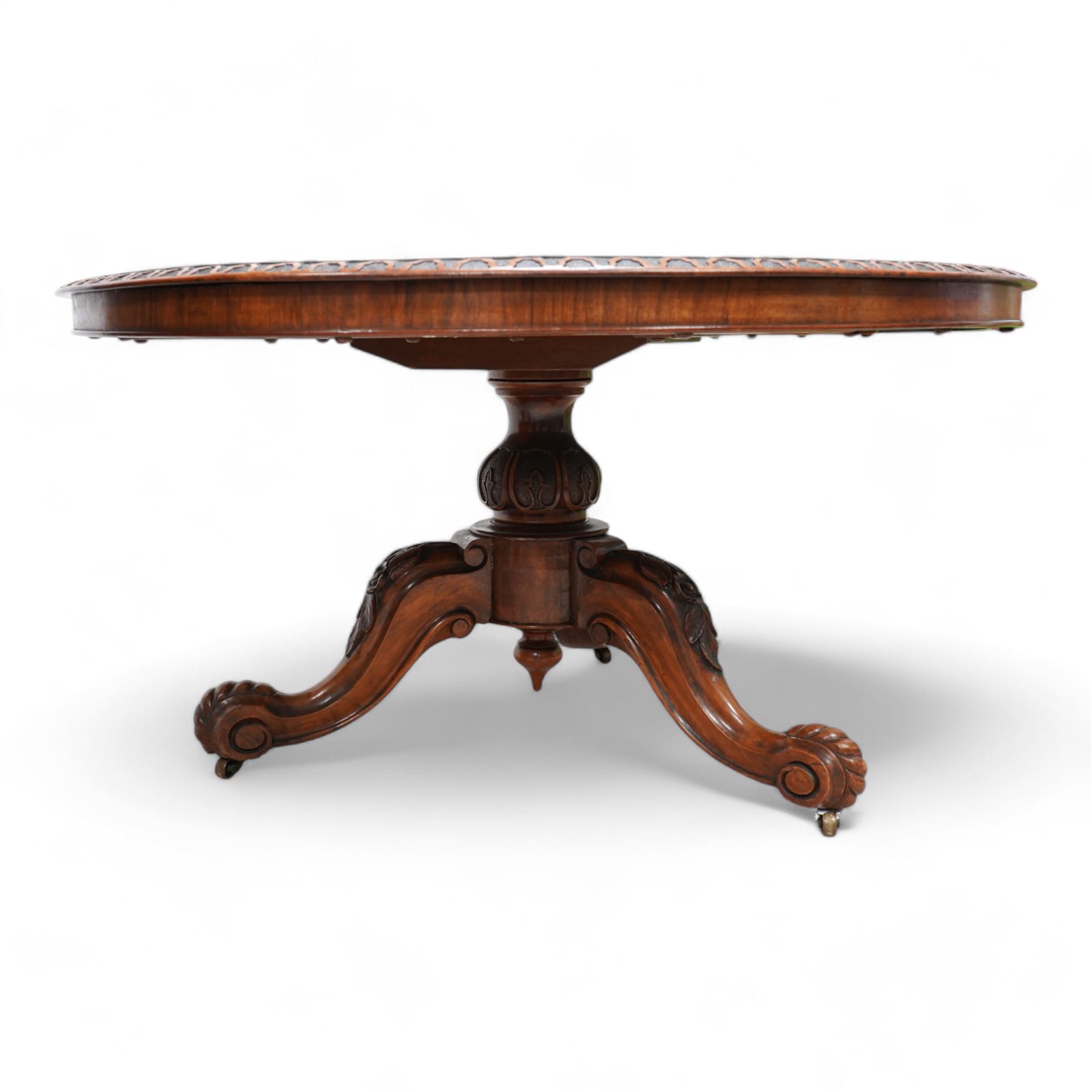 Victorian figured walnut breakfast or centre table, circular tilt-top with carved edge and quarter-matched veneers, turned pedestal with foliate carved baluster on three out splayed supports decorated with floral carvings and scrolled terminals, on brass castors 