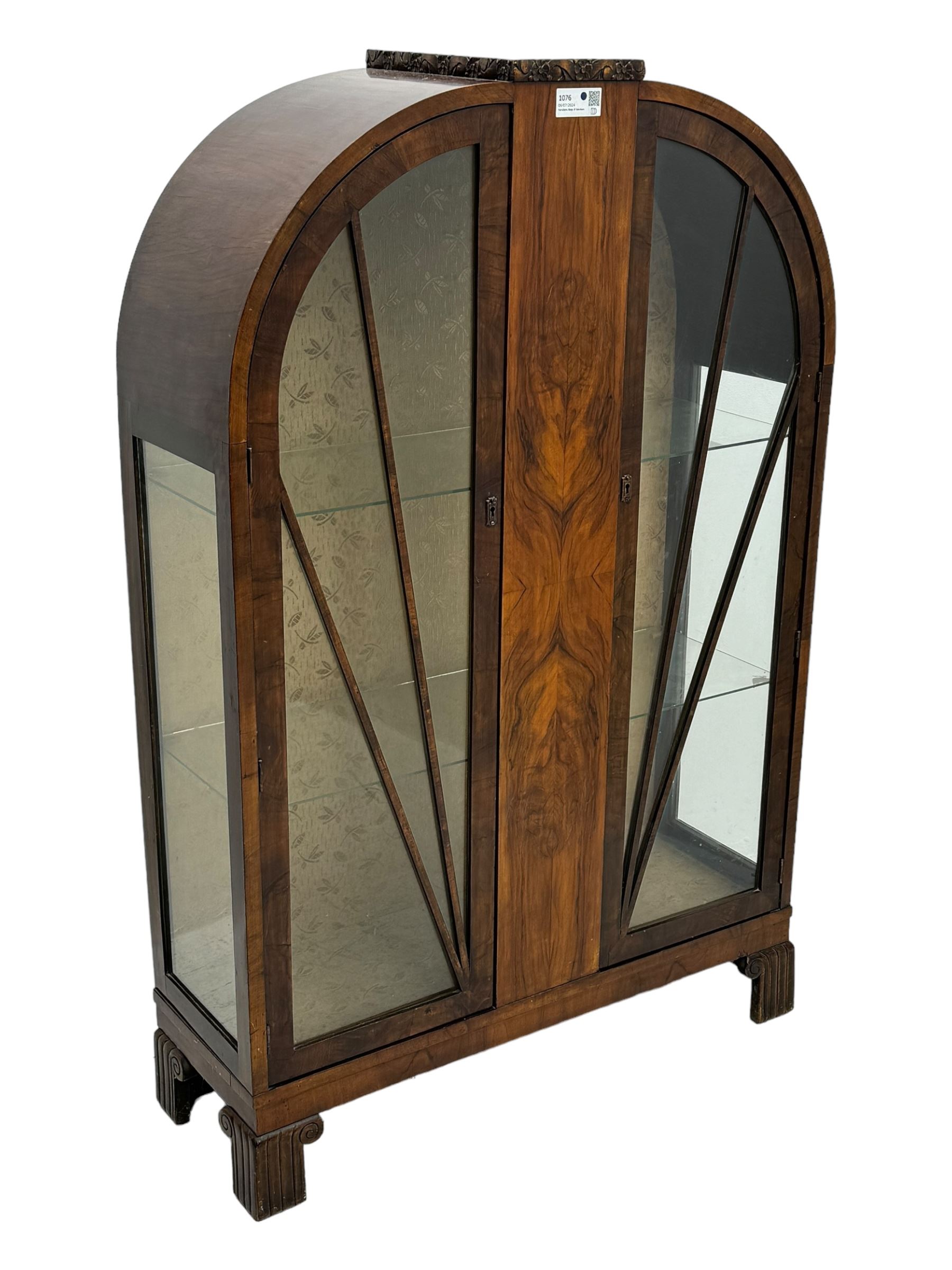 Early 20th century Art Deco walnut display cabinet, with arched top and carved floral crest, central band flanked by two glazed panel doors with decorative mullions, enclosing two glass shelves, on scroll carved bracket feet