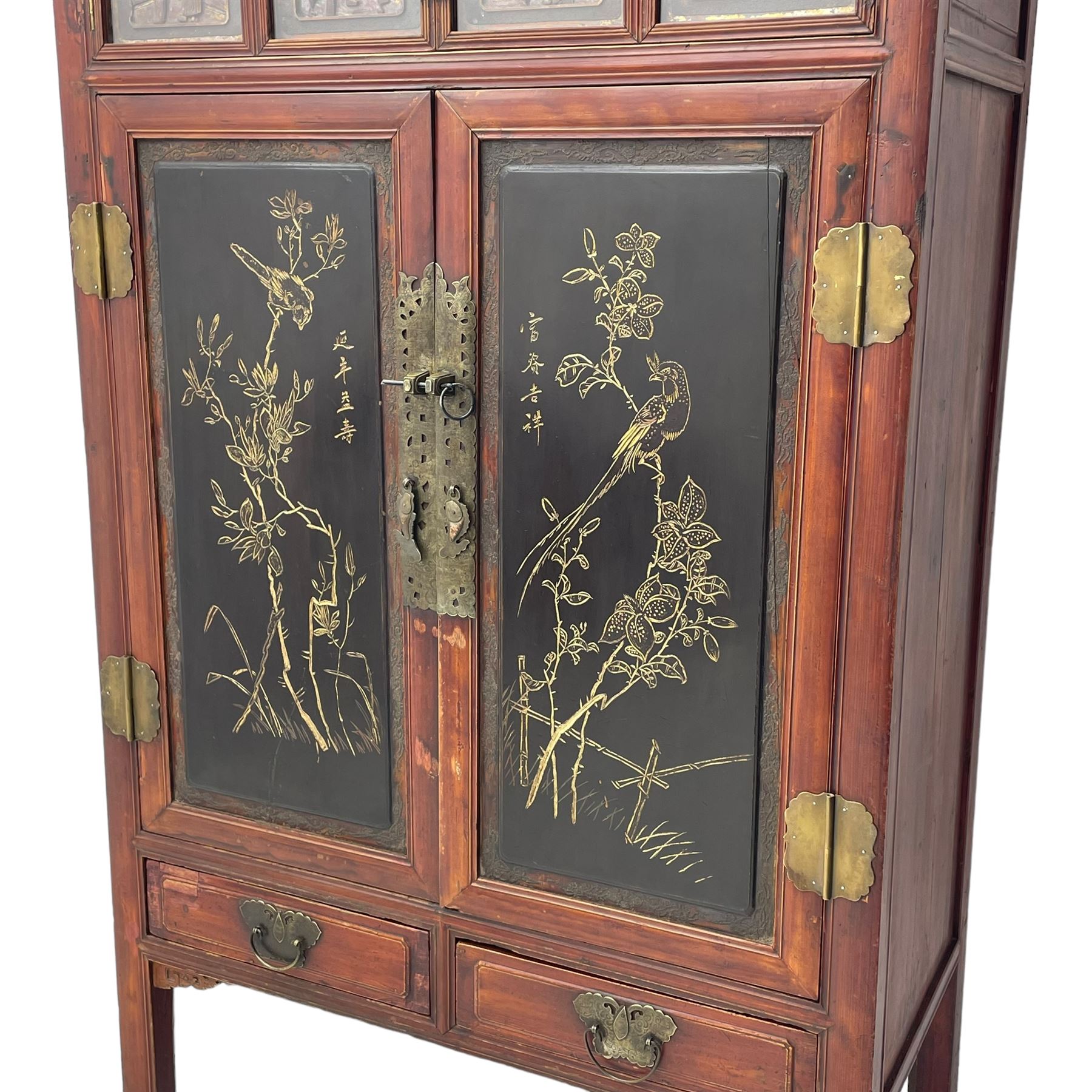 Late 19th century Chinese Qing dynasty red and black lacquered wood cabinet, Fujianese province, upper cupboard enclosed by two doors with relief carved and gilt panels depicting figures behind glass, the large cupboard enclosed by two panelled doors carved with trailing foliate branches and birds, inscribed with Chinese characters, to the left ‘Prolong life’ and to the right ‘Wealth and good fortune’, fitted with two drawers, square supports with carved brackets 