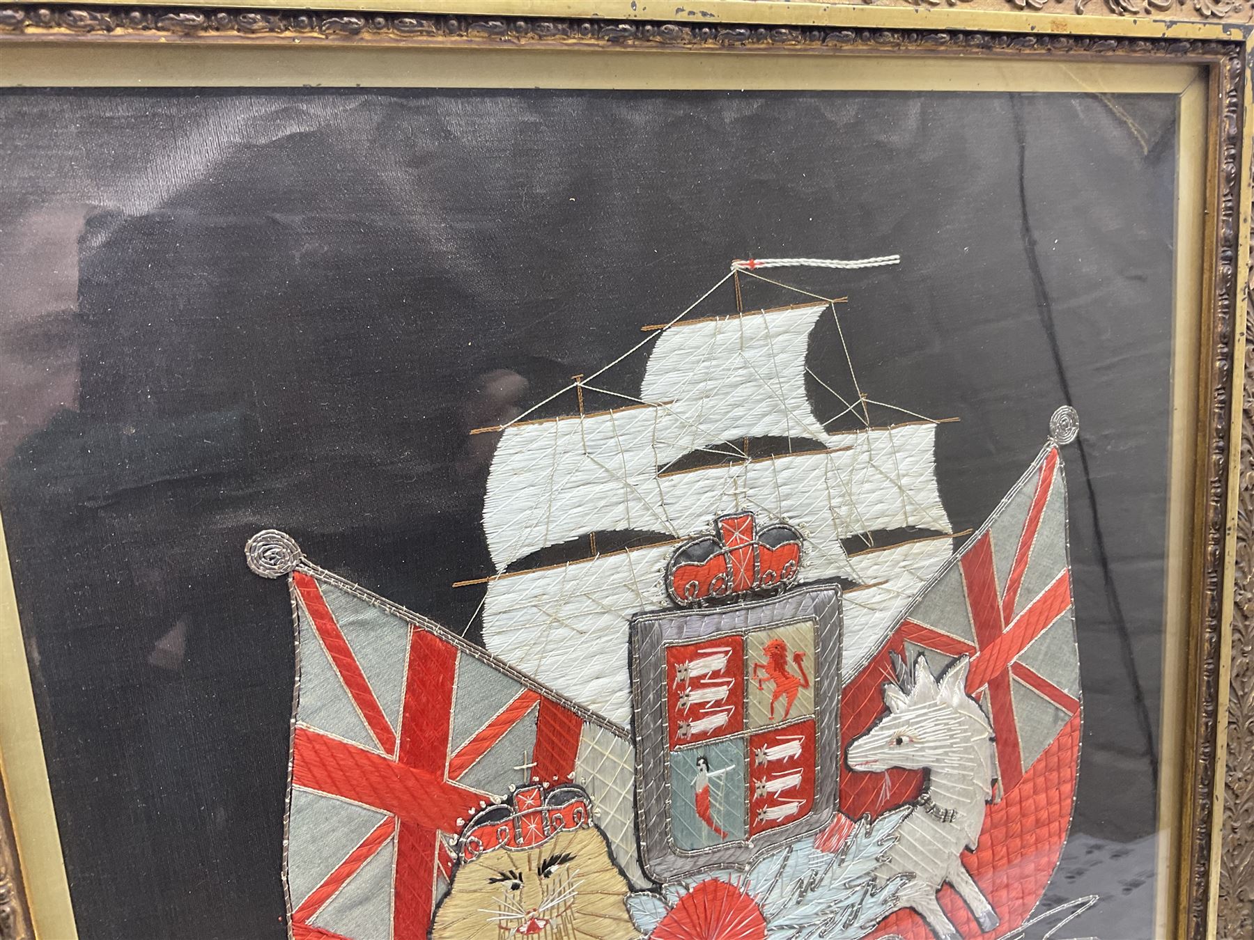 Early 20th century silk needlework panel, depicting a ship in full sail and Royal Coat of Arms to centre, with red and white ensigns to either side, lion and unicorn below with quote 'Dieu Et Mon Droit', upon a black silk ground within gilt frame, H59cm