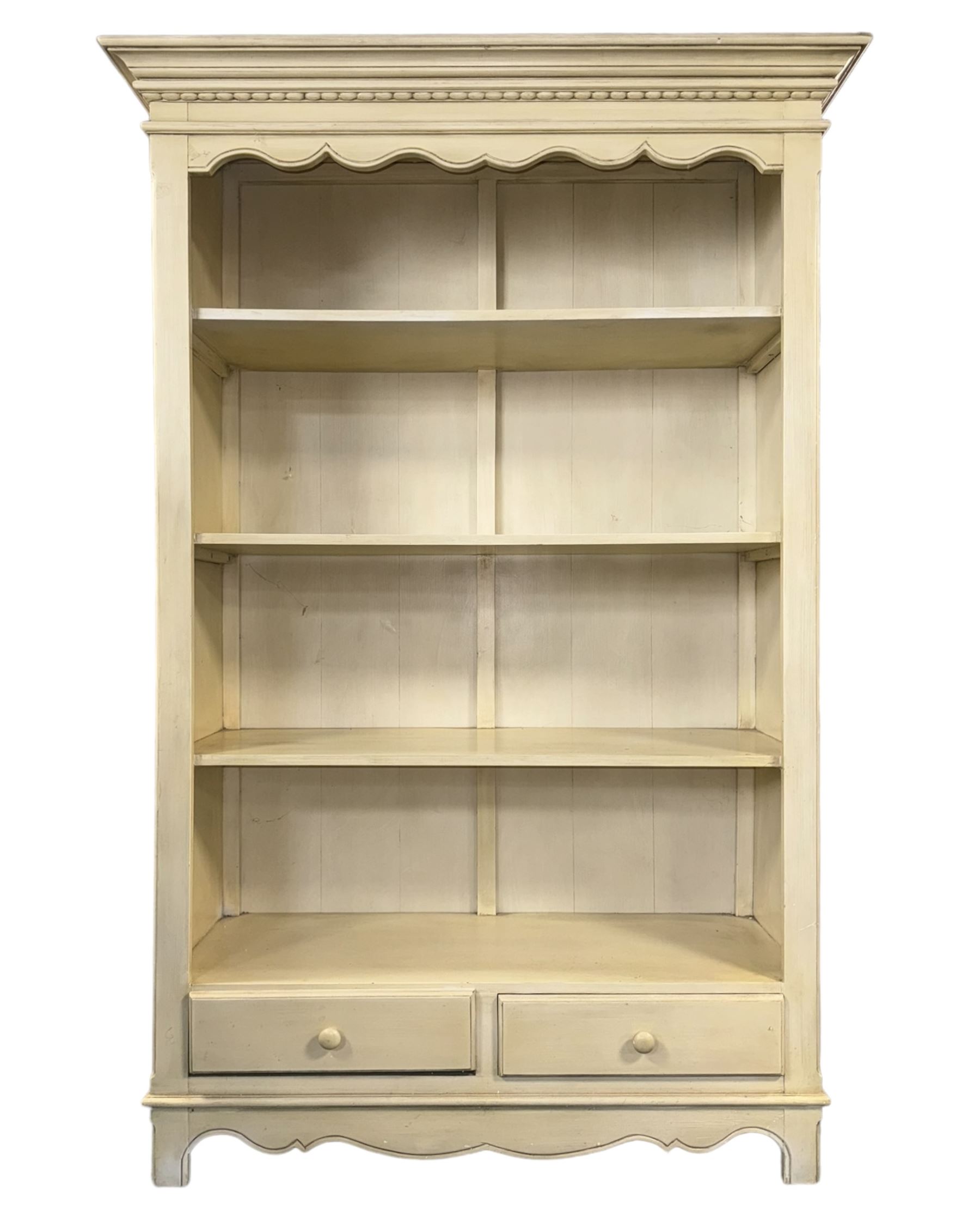 Provincial style cream painted bookcase, projecting cornice over dentil moulding, scalloped frieze above three open shelves, lower section fitted with two drawers, on bracket feet with apron