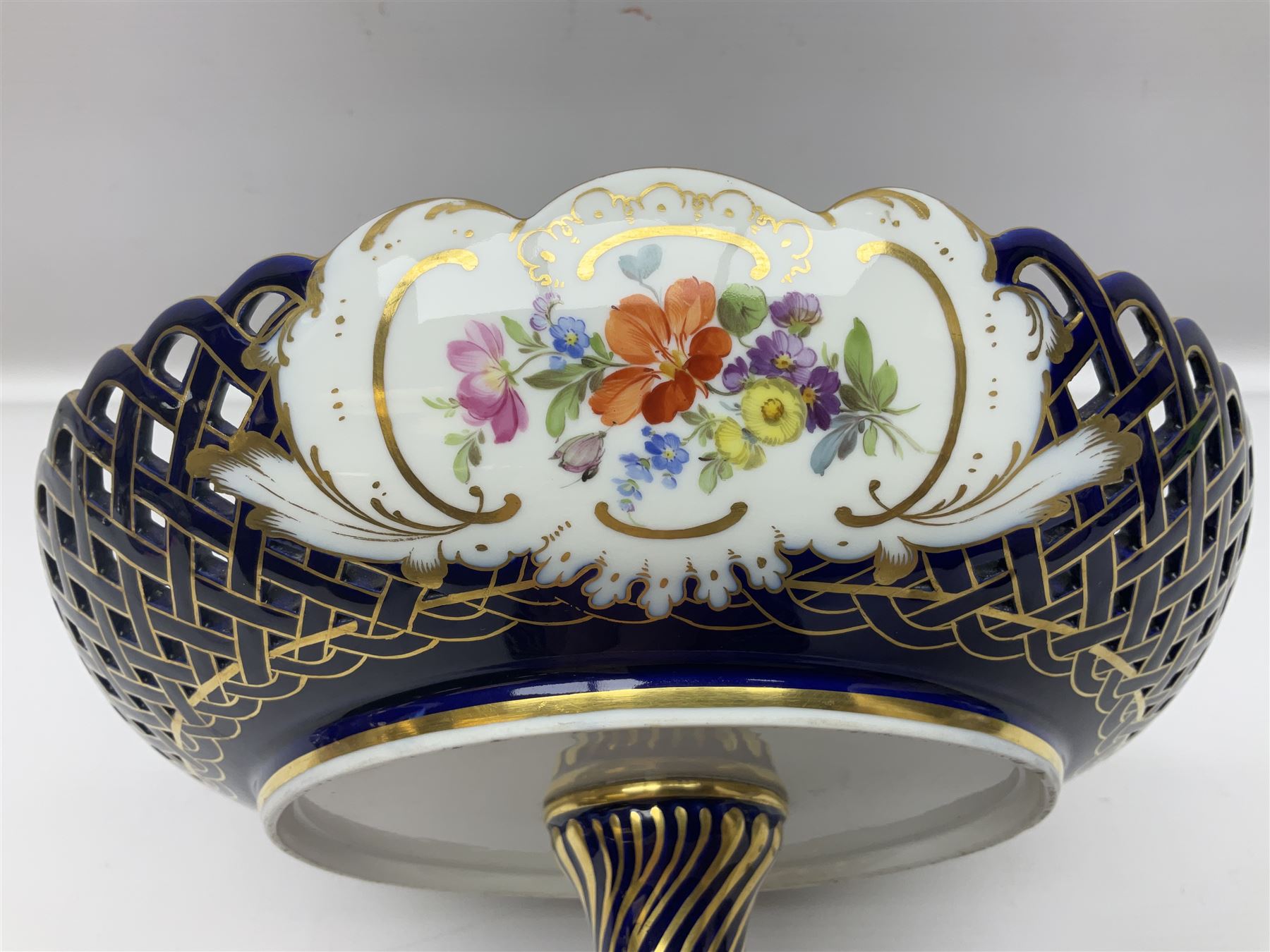 Meissen style pedestal dish, with central panel depicting a trading scene in a harbour setting, the bowl with openwork lattice sides divided by three hand painted floral panels, upon a blue ground with gilt detailing, upon a fluted knopped pedestal and circular foot, H22cm