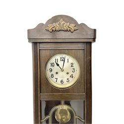 Oak cased 1930s 8-day German wall clock - with a crested top, glazed door and visible pendulum, silvered dial with Arabic numerals and spade hands, two train movement striking the hours on a gong. With pendulum and key.