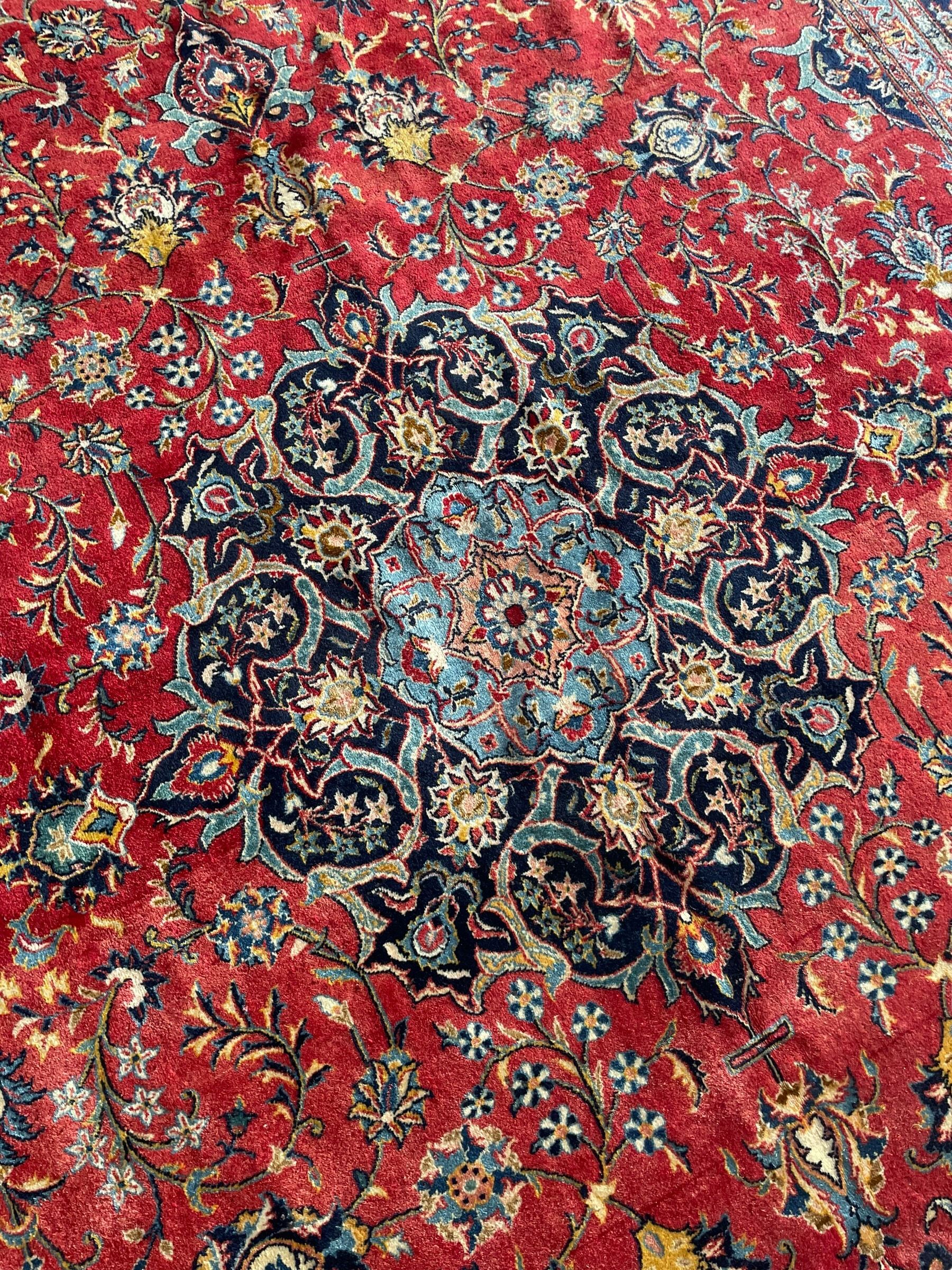 Persian Kashan crimson ground carpet, central rosette medallion on a field of trailing foliate branches decorated with palmettes, scrolling border with repeating plant motifs and flower heads, enclosed within floral pattern guard stripes