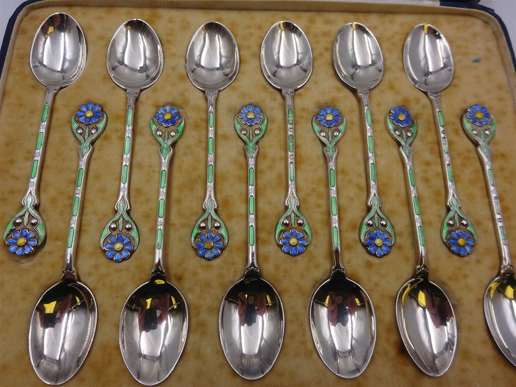 Set of twelve silver coffee spoons, each with enamelled blue flower to terminal and green and white enamel decoration to stem, hallmarked Mappin & Webb Ltd, Birmingham 1934, contained within fitted case