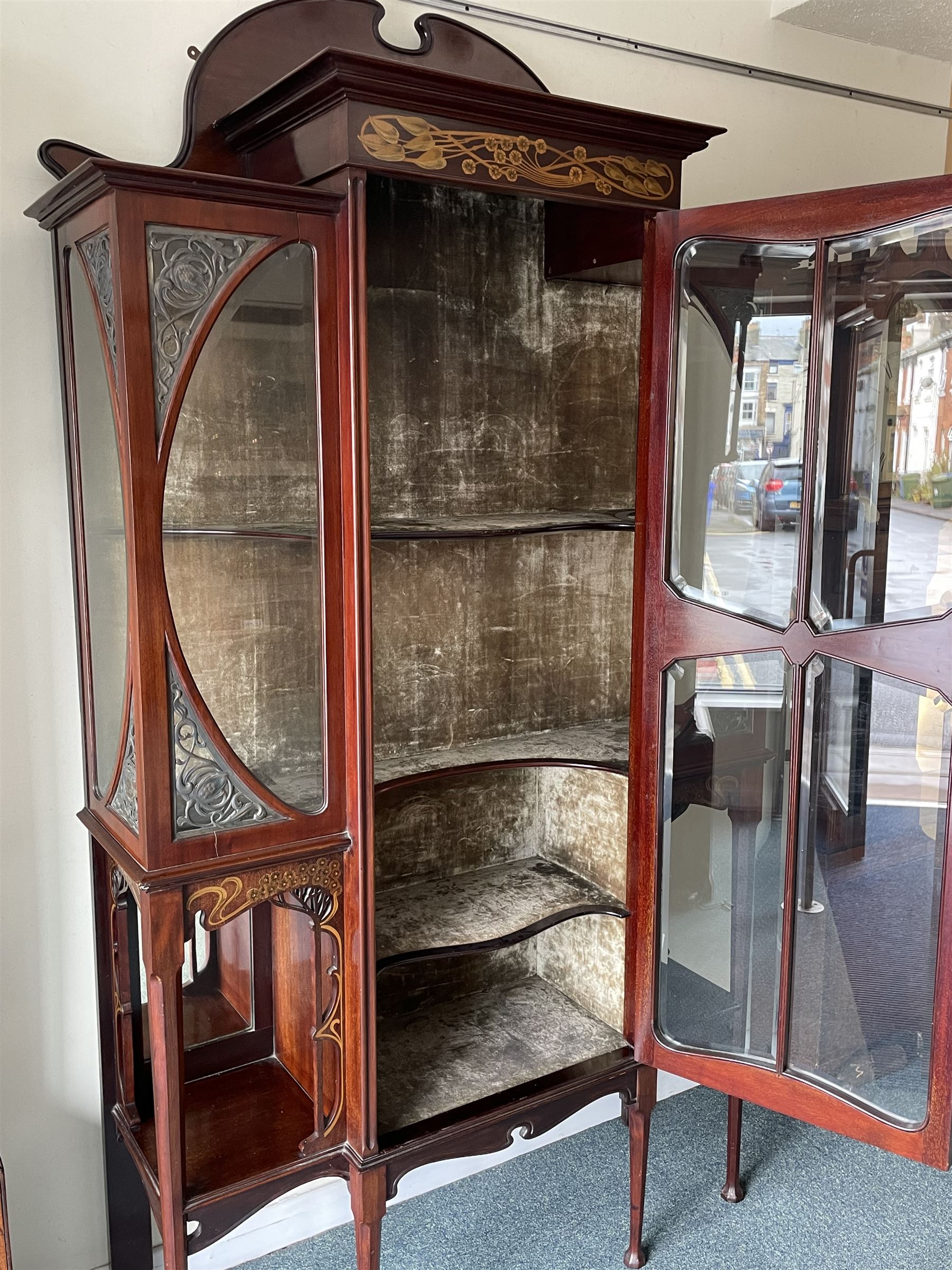 Art Nouveau period inlaid mahogany display cabinet, raised shaped and moulded pediment over projecting moulded cornices, the upper frieze inlaid with trailing branches and tulips, single astragal and bevel glazed door enclosed velvet lined interior fitted with shelves, flanked by curved glass panes decorated with pierced metal foliate spandrels, lower flanking shelves with mirror backs enclosed within shaped and pierced rails inlaid with flower heads, on tapering octagonal supports terminating to compressed pad feet, the lock stamped 'Salmon Bros, London' 