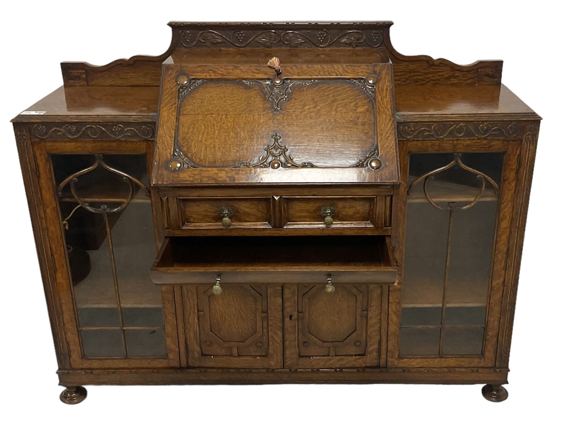 Early 20th century heavily carved oak bookcase bureau, raised back with grape-vine carved frieze, over fall-front enclosing fitted interior, above two short drawers and curved frieze drawer, flanked by astragal glazed and panelled cupboards, on turned feet