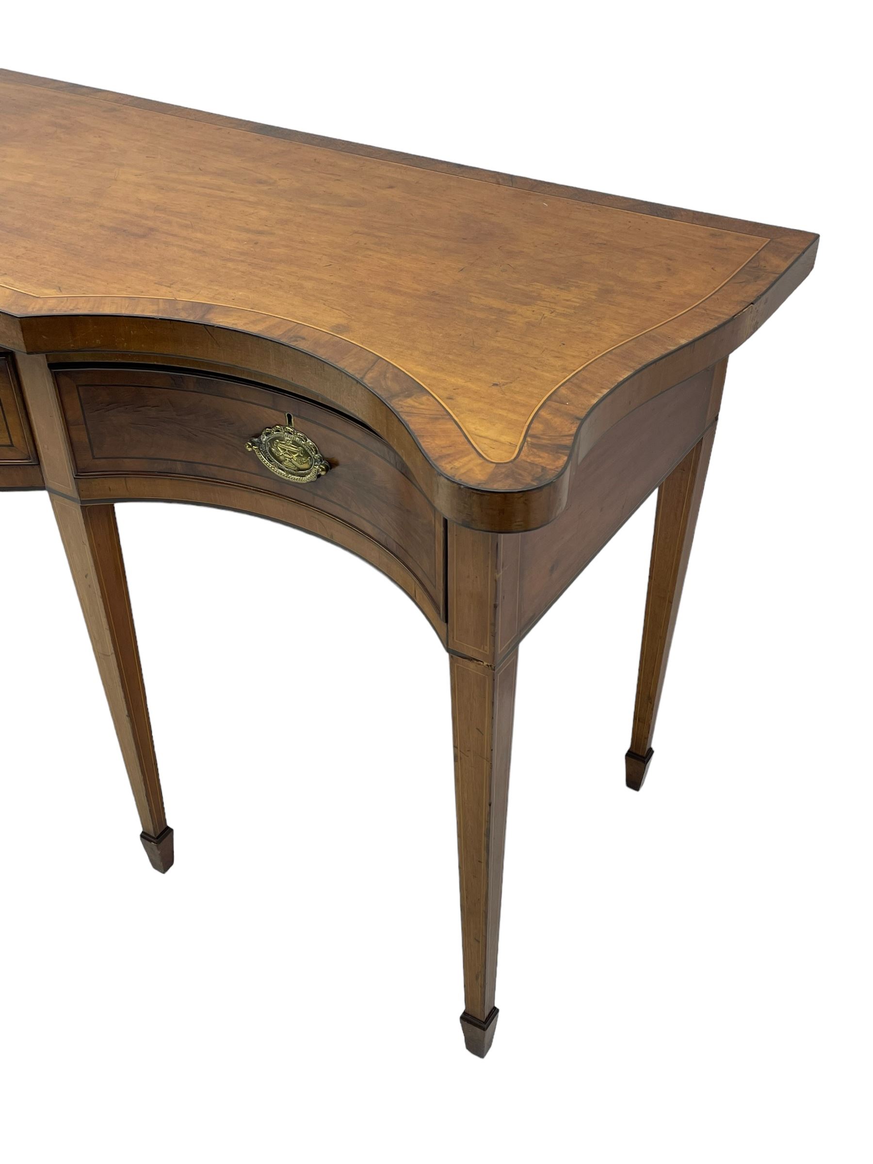 Large George III mahogany serpentine serving table, shaped top with crossing banding and stringing, the frieze fitted with three cock-beaded drawers, oval pressed brass handle plates decorated with urns, on square tapering supports with spade feet 