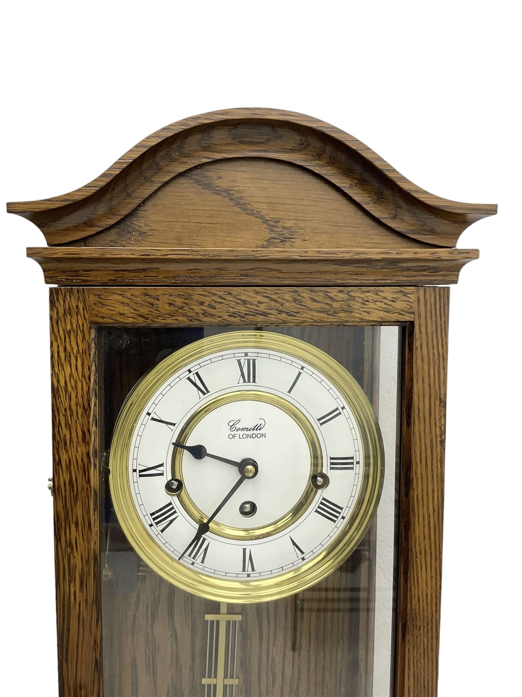 Commiti of London -  three train spring driven 8-day wall clock in an oak case, with a fully glazed door and curved pediment, two part dial with Roman numerals, spade hands and gridiron pendulum, rack movement chiming the hours and quarters on 5 gong rods. With key.