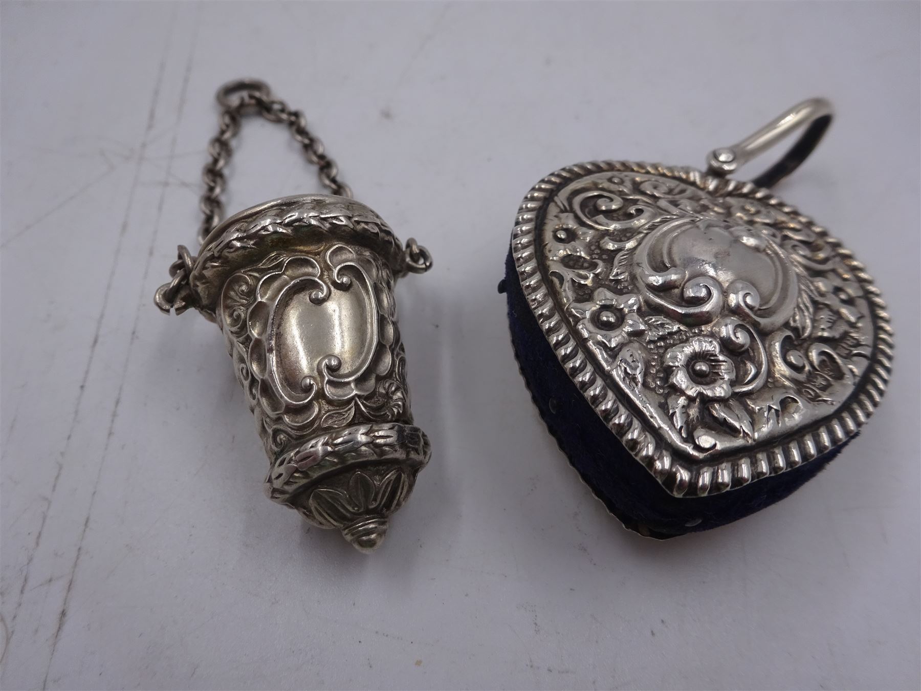 Edwardian silver pin cushion, in the form of a heart, with embossed floral decoration, with blue pin cushion between two heart shaped silver covers, with chatelain hook, hallmarked Henry Matthews, Birmingham 1902, together with a late Victorian silver thimble holder, embossed with scrolling decoration and with velvet interior, with chatelain chain, hallmarked Levi & Salaman, Birmingham 1899, pin cushion H4.5cm