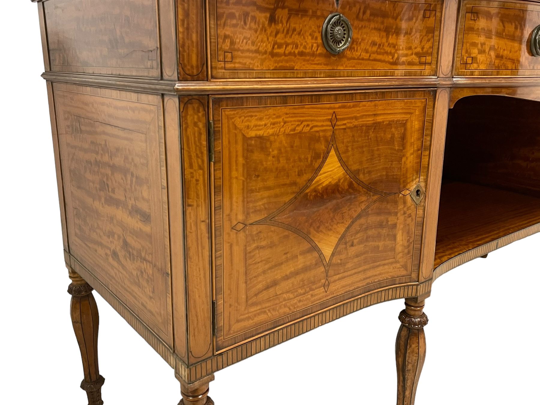 Waring & Gillow - Edwardian Sheraton Revival satinwood sideboard, shaped and stepped break bow-front, the top with central oval book-matched veneered panel and rosewood banding, the front and return edges inlaid with a single row of repeating circles, three frieze drawers each inlaid with rosewood band and ebony stringing, two cupboards flanking open centre, the doors inlaid with lozenges and satinwood cross bandings, on faceted octagonal supports with foliate carved collars, terminating to turned feet, the central drawer with 'Warings' metal plaque and stamped 'Gillows'