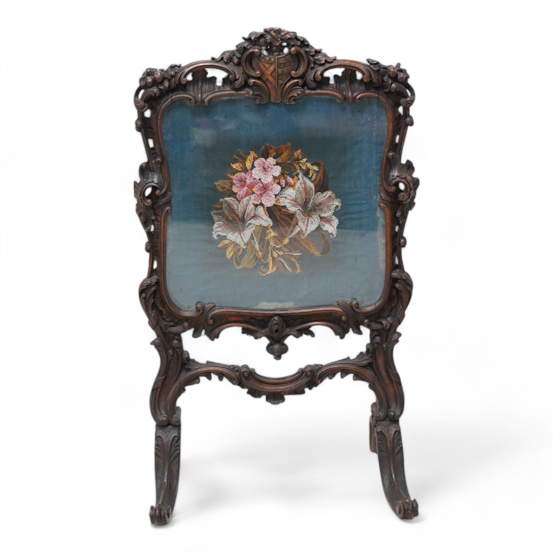 19th century carved wood fire screen, shield pediment decorated with oak leaves and acorns, the surrounding shell carved frame decorated with scrolling foliage, bead needle work panel depicting floral bouquet, on foliate carved splayed supports with scrolled terminals 