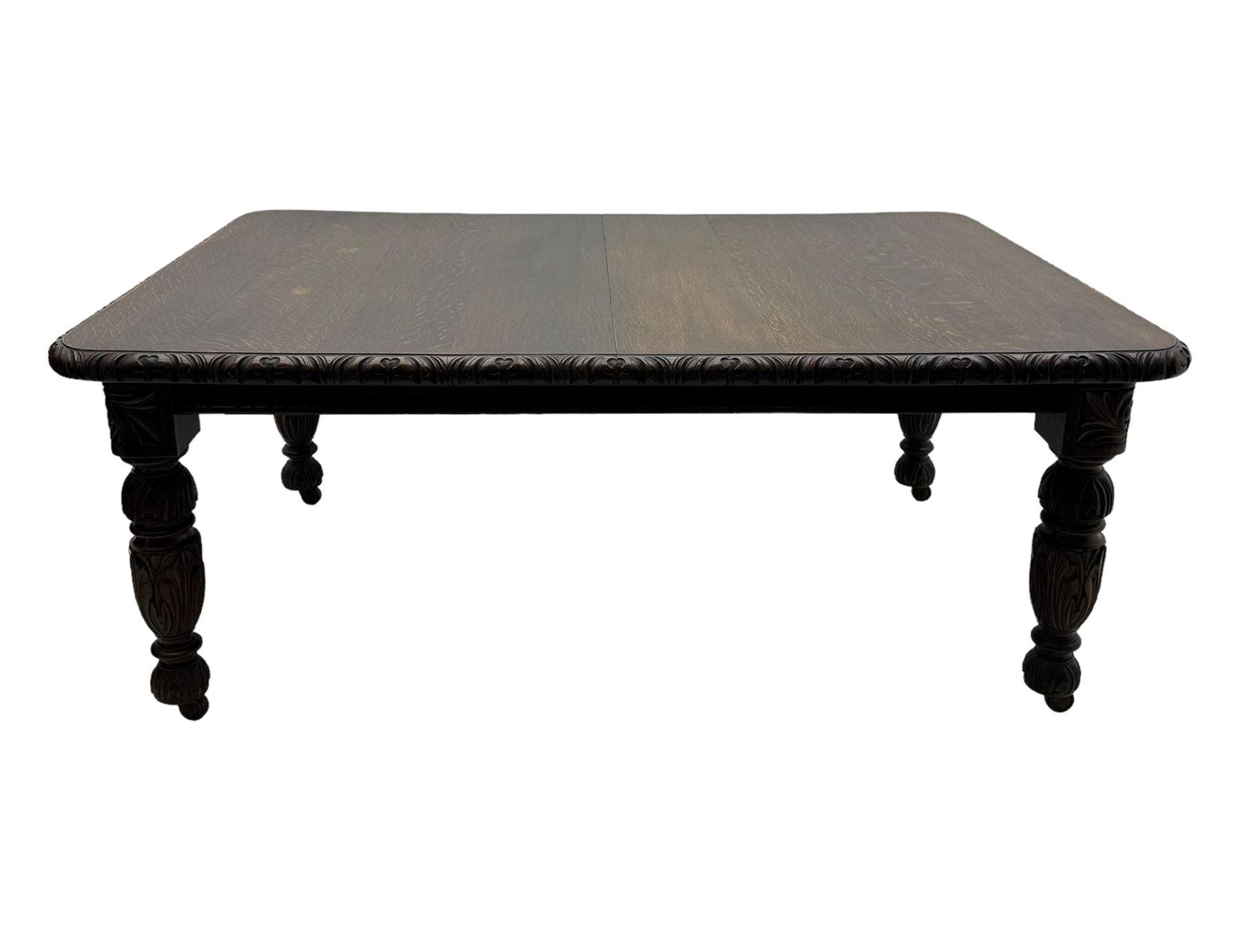 19th century heavily carved oak extending dining table, rectangular top with rounded corners and carved gadrooned edge, extending via winding mechanism with two additional leaves, raised on acanthus leaf-carved baluster supports terminating in ceramic castors