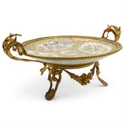 19th century Chinese Canton famille rose circular dish, painted with figures and exotic birds in shaped panels, against a floral and scroll ground, mounted in ormolu style gilt metal twin handled mount with beaded border and three scroll feet, W30cm x H14cm