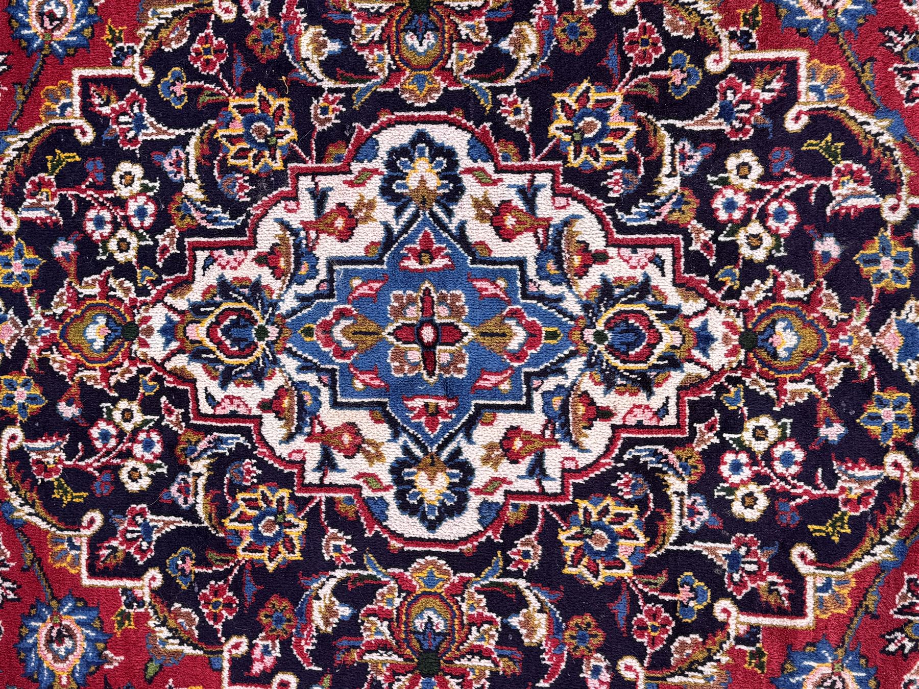 Large Kashan crimson ground carpet, central indigo medallion with stylised floral motifs, flanked by matching spandrels, bordered by multiple bands featuring repeating foliate and floral designs throughout