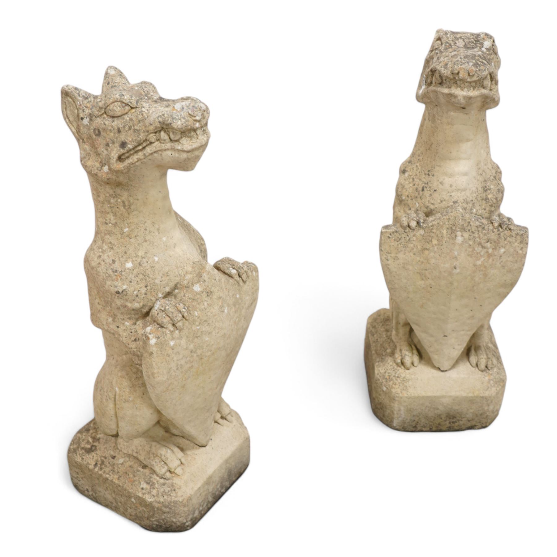 Near pair of cast stone garden seated dragons, snarling jaw holding shield on chamfered base 