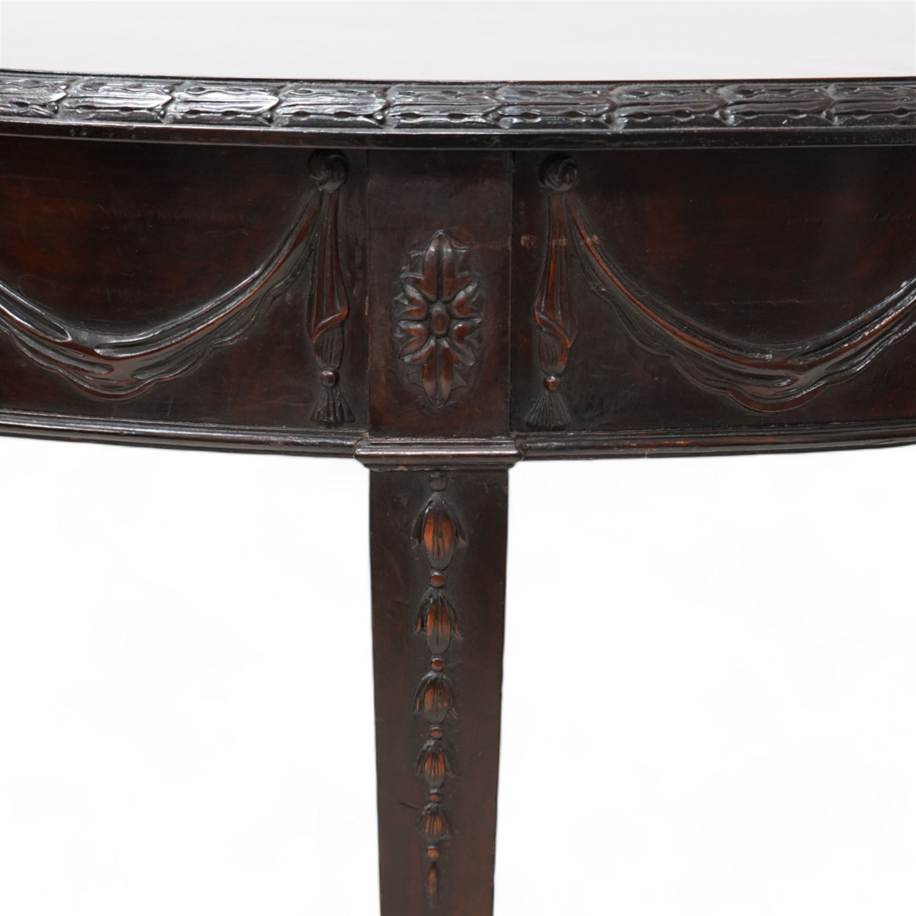 Early 20th century Hepplewhite Revival mahogany console table, demi-lune top with carved moulded edge, the frieze decorated with moulded repeating festoons, raised on square tapering supports with applied bell-flower motifs, terminating in spade feet