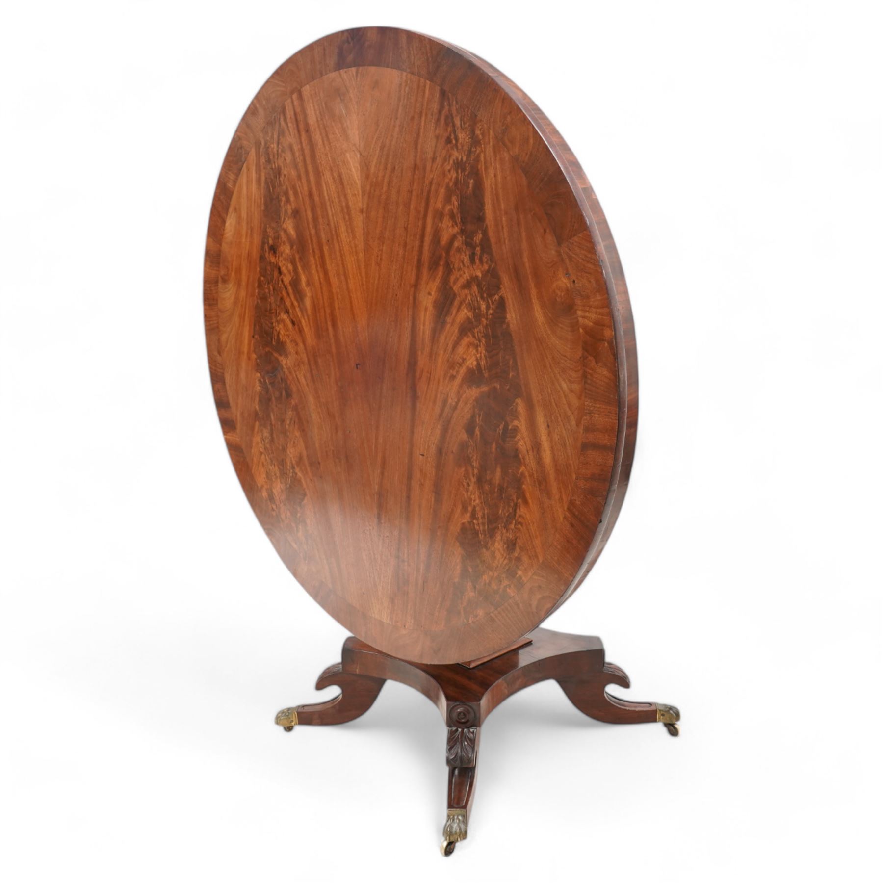 William IV mahogany breakfast or centre table, circular tilt-top with book-matched figured veneers within crossbanding, tapered square column on concaved square platform, four splayed acanthus carved feet with brass hairy paw castors 