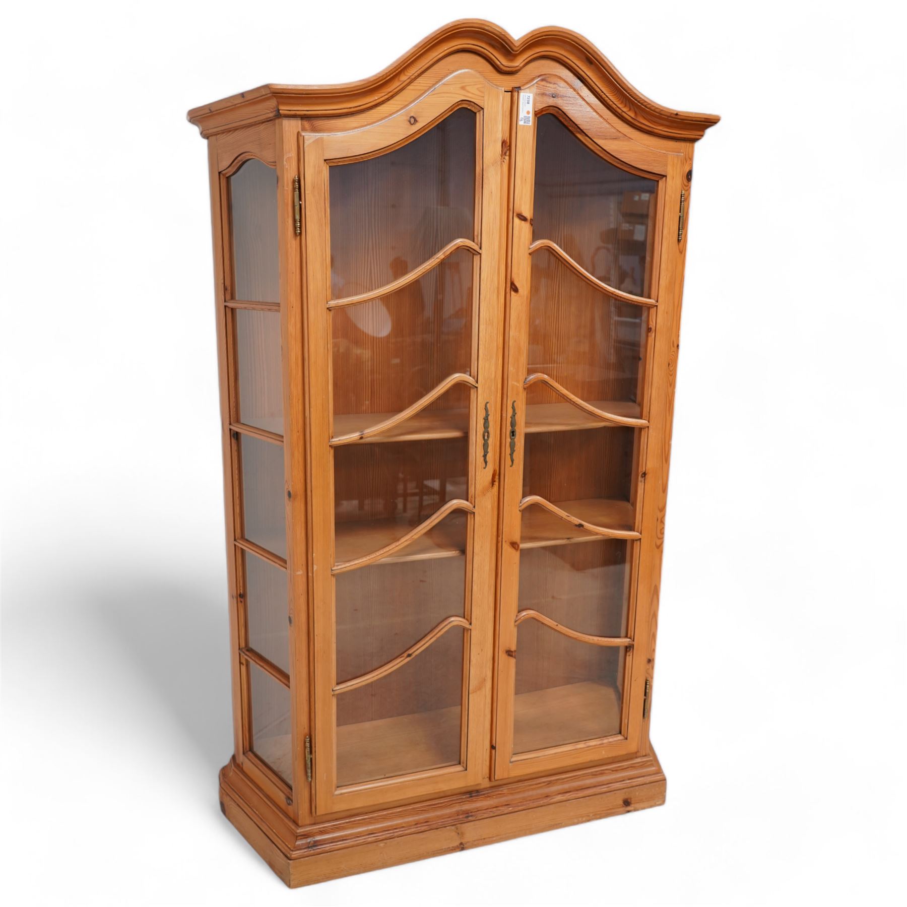 Polished pine display cabinet, shaped top over two glazed doors, fitted with three shelves, on moulded plinth base 