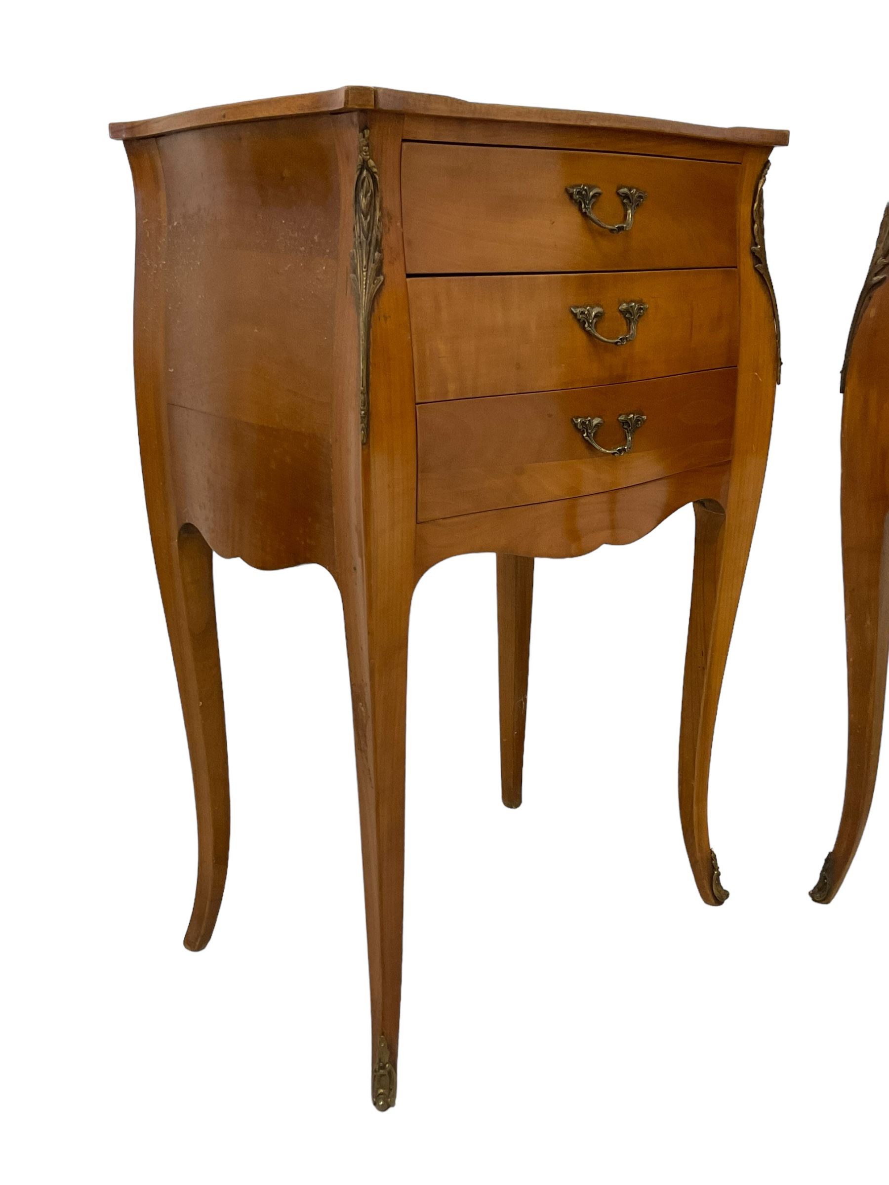 Pair of late 20th century French design cherry wood bedside lamp tables, shaped bombe form, fitted with three drawers, on tapering cabriole supports, decorated with ornate cast gilt metal mounts 