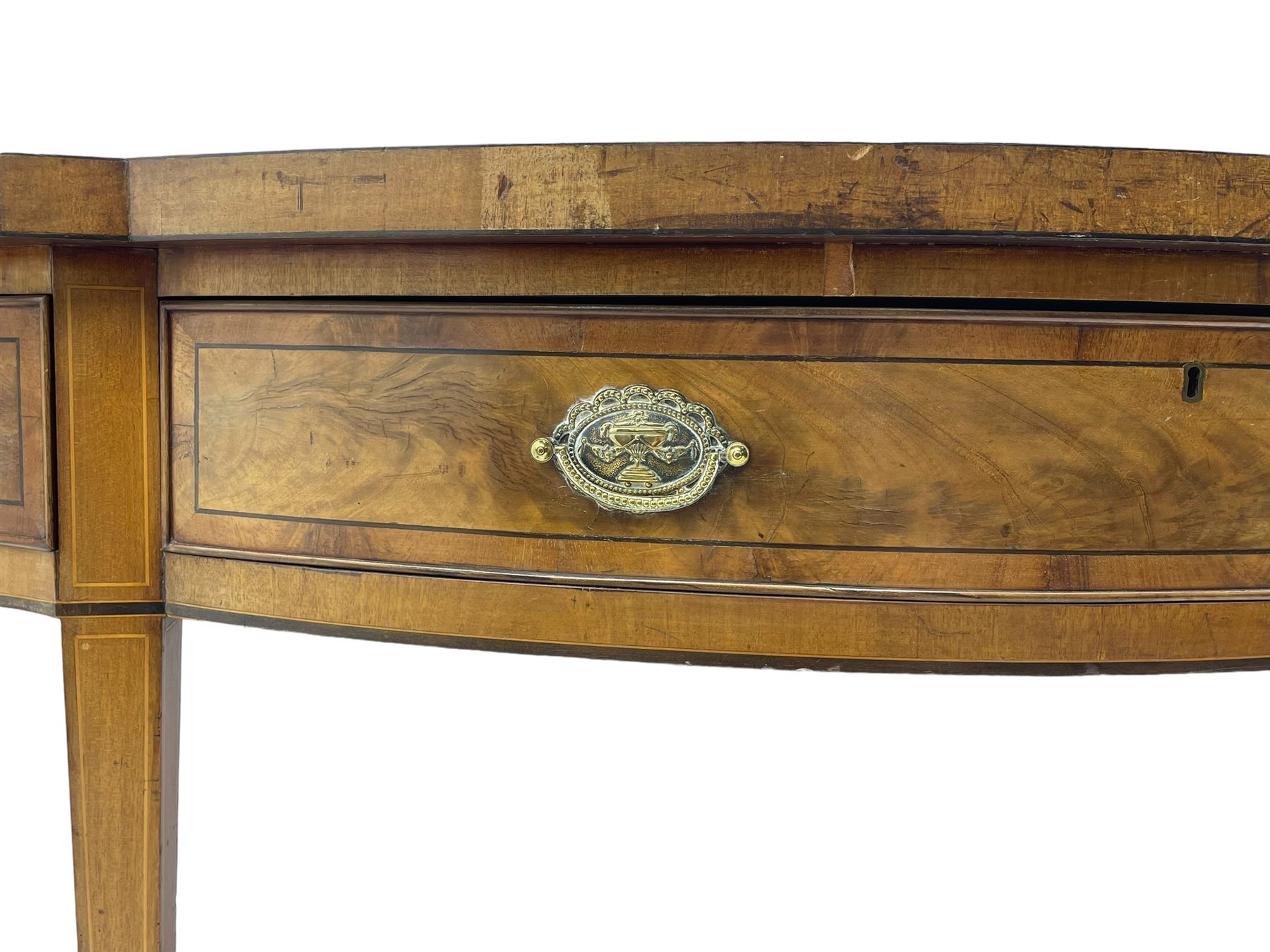 Large George III mahogany serpentine serving table, shaped top with crossing banding and stringing, the frieze fitted with three cock-beaded drawers, oval pressed brass handle plates decorated with urns, on square tapering supports with spade feet 