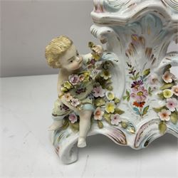 Early 20th century porcelain garniture, the central shaped basket upon a column supported by a female figure, and base with scroll feet, flanked by a pair of four branch candelabra, the scrolling branches and central stem leading to urn shaped sockets over foliate drip pans, upon shaped columns and bases with applied putto, the whole with encrusted flowers throughout, basket H33.5cm, candelabra H52cm