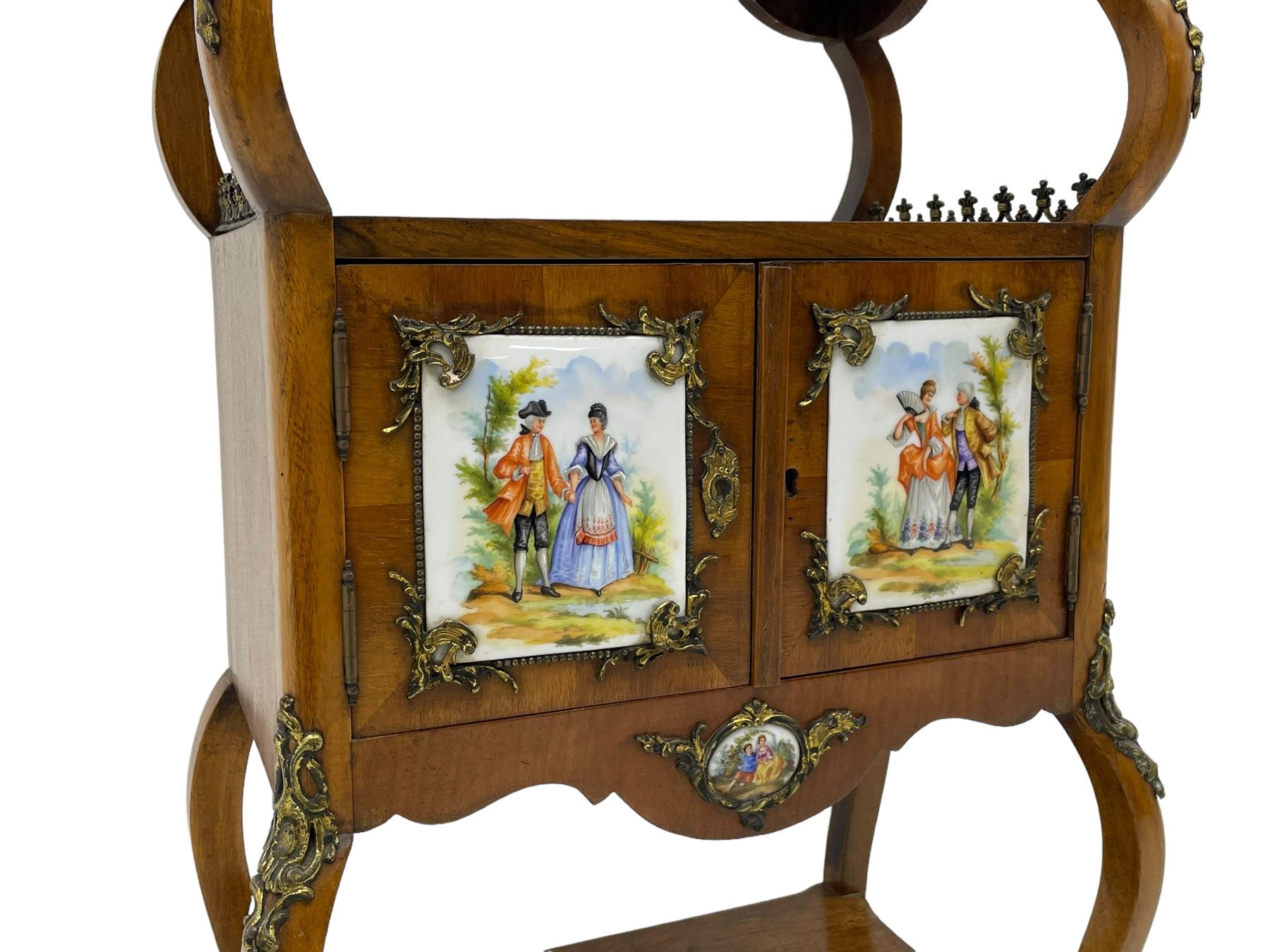 Mid-to-late 20th century French figured walnut three-tier bedside stand, shaped moulded top inlaid with figured book-matched veneers and crossbanding, double cupboard below enclosed by two doors mounted by painted porcelain panels depicting courting scenes within landscapes, on cabriole supports united by undertier, decorated with ornate cast metal cartouche mounts 