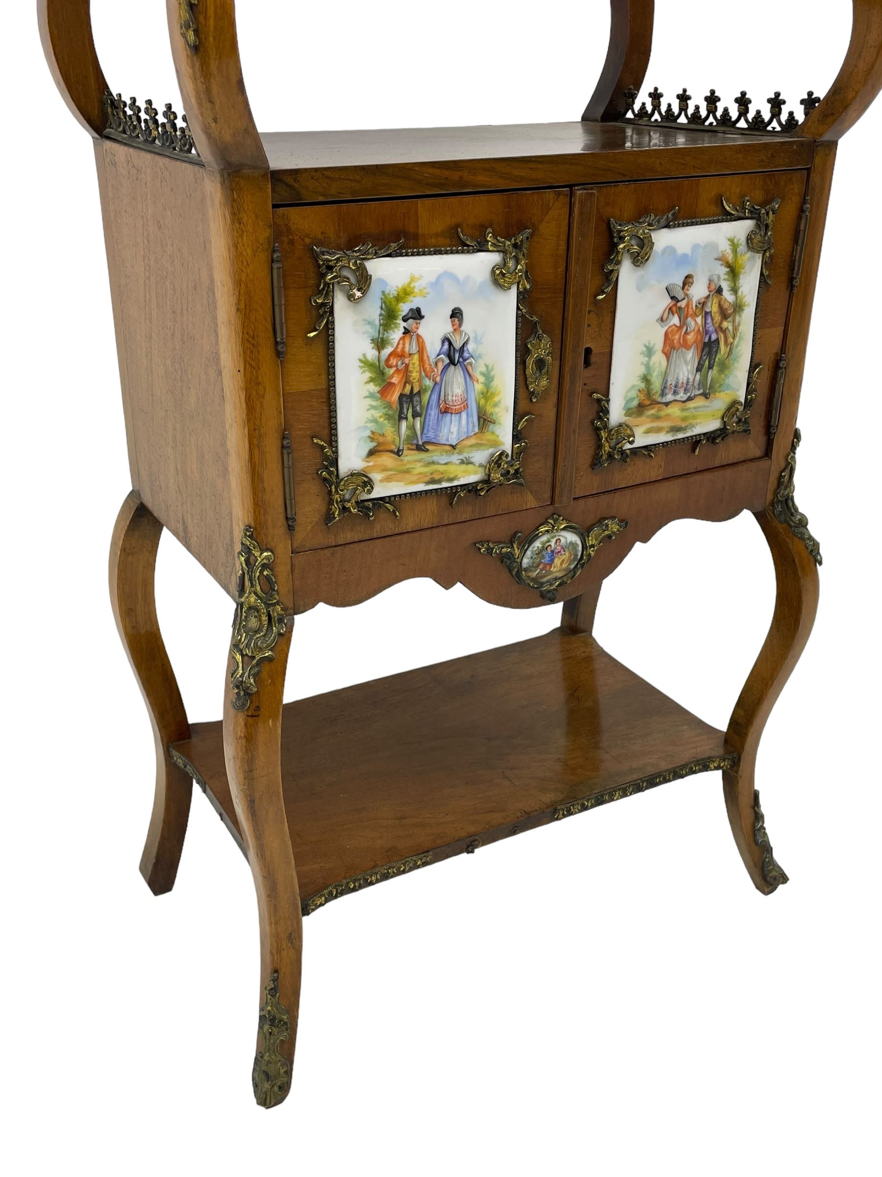 Mid-to-late 20th century French figured walnut three-tier bedside stand, shaped moulded top inlaid with figured book-matched veneers and crossbanding, double cupboard below enclosed by two doors mounted by painted porcelain panels depicting courting scenes within landscapes, on cabriole supports united by undertier, decorated with ornate cast metal cartouche mounts 