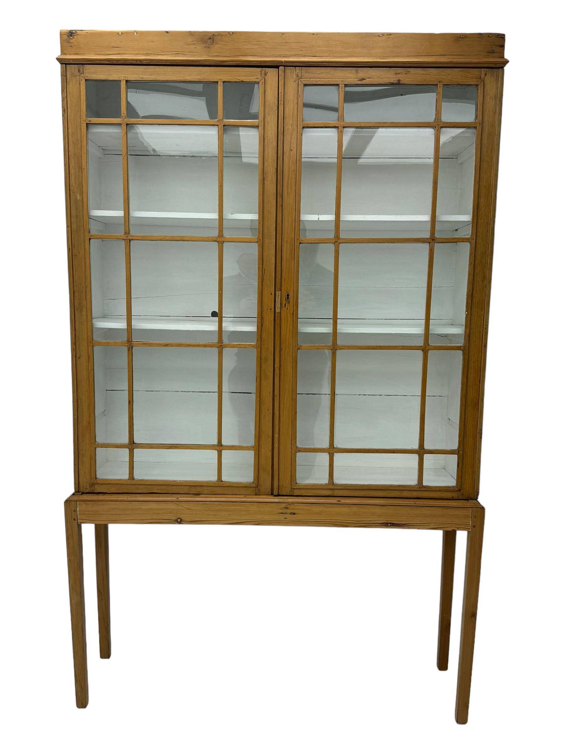 Georgian stripped pine bookcase display cabinet, plain moulded cornice over two glass-panel doors with multiple panes, enclosing white painted interior with three shelves, raised on square supports