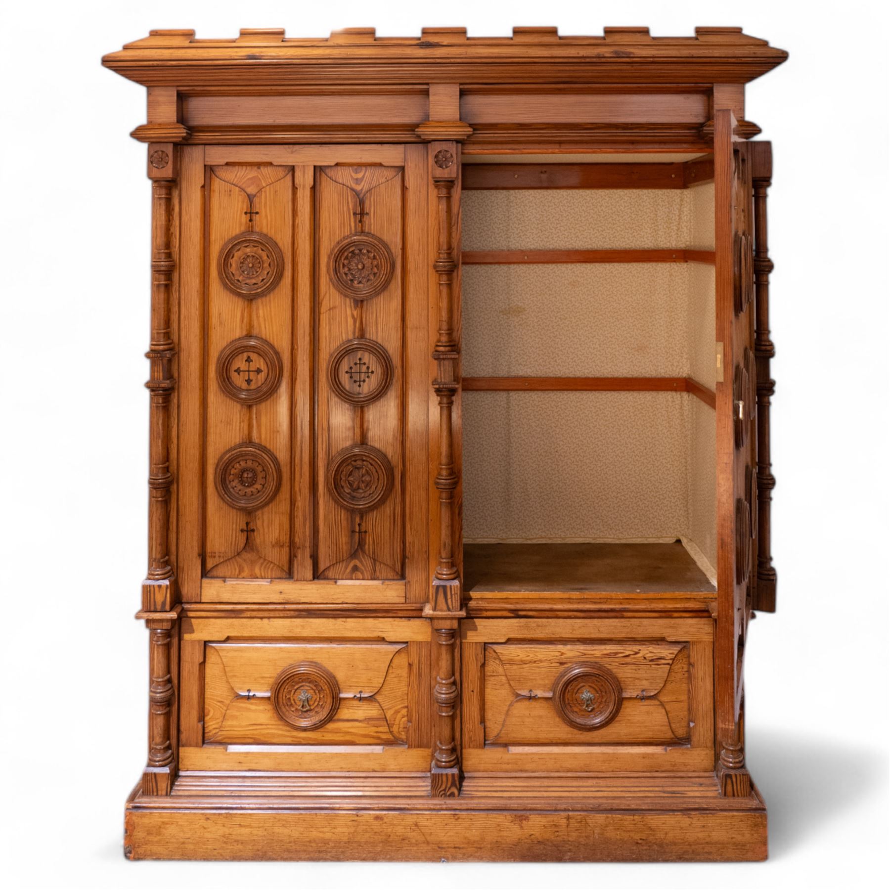 19th century pitch pine ecclesiastical vestment wardrobe or sacristy cabinet, crenelated and projecting moulded cornice over two panelled doors, the panelled doors carved with linen folds and mounted with rows of roundels depicting crucifixes and flower heads, enclosed by turned pilasters, two drawers to the base, on moulded plinth base 