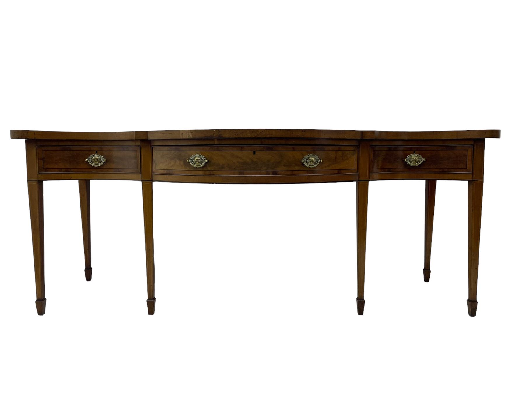 Large George III mahogany serpentine serving table, shaped top with crossing banding and stringing, the frieze fitted with three cock-beaded drawers, oval pressed brass handle plates decorated with urns, on square tapering supports with spade feet 