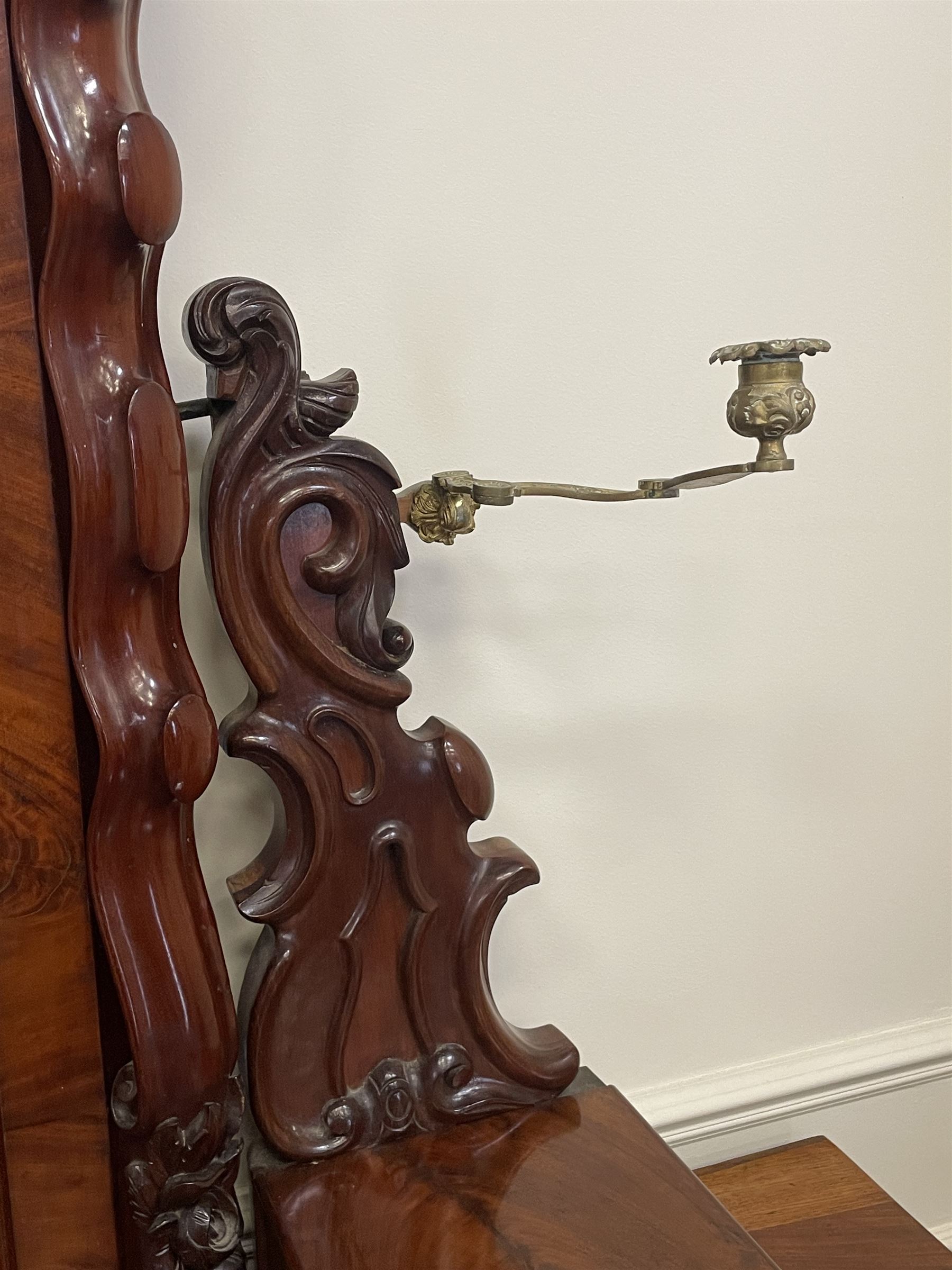 Tall Victorian figured mahogany twin pedestal dressing table, the tall mirror with flower head and scrolled leaf carved pediment, flanked by two turned and carved finials over faceted lancet arched lantern tops, plane mirror plate enclosed by shaped frame, pierced and undulating moulded uprights, on C-scroll and curled leaf carved supports fitted with cast brass hinged candle sconces, each pedestal fitted with two small trinket drawers over larger drawer and panelled cupboard, canted corners with matched upright mounts, the cupboard doors enclosing slides, on scroll carved feet 