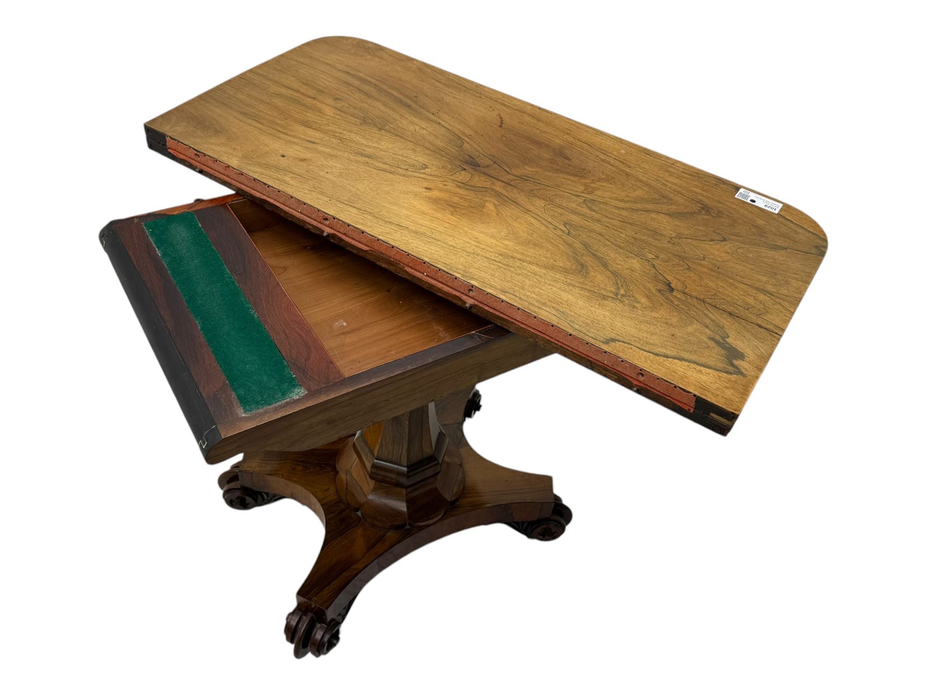 Early Victorian rosewood card table, rectangular foldover swivel top with rounded corners, red baize-lined surface, frieze adorned with carved foliate motifs, supported by a central octagonal waisted column on shaped quatrefoil base, terminating in scrolled feet with concealed metal castors