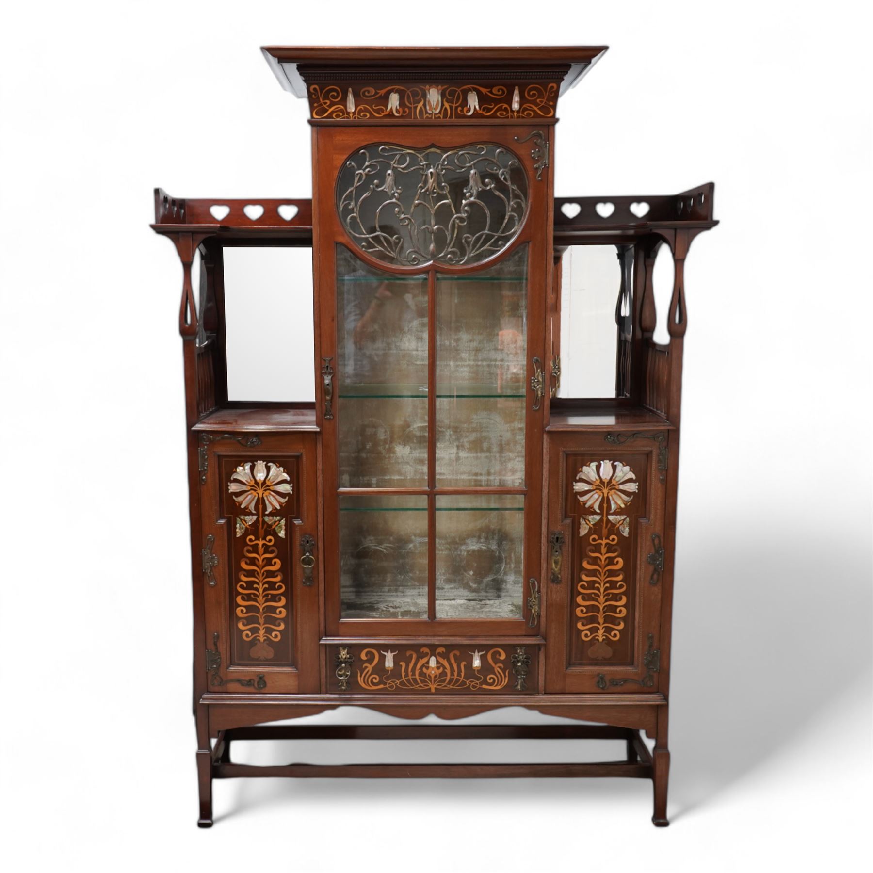 Shapland & Petter; Barnstaple (1854-1999) - Art Nouveau circa. 1900s 'Lily' mahogany display cabinet, raised top with projecting moulded cornice, the frieze inlaid with flower heads and scrolling branches in boxwood and mother of pearl, enclosed by central bevel glazed door mounted with open scrolling metalwork, flanking bevelled mirror plates over panelled cupboards, the panels inlaid with fanned lily heads over butterflies and scrolled tapering leaves, the upper gallery pierced with heart motifs over swelled pierced supports and balustrades, single central drawer with matching inlay, on square tapering and block feet united by plain stretchers, the locks stamped 'S&B, B'