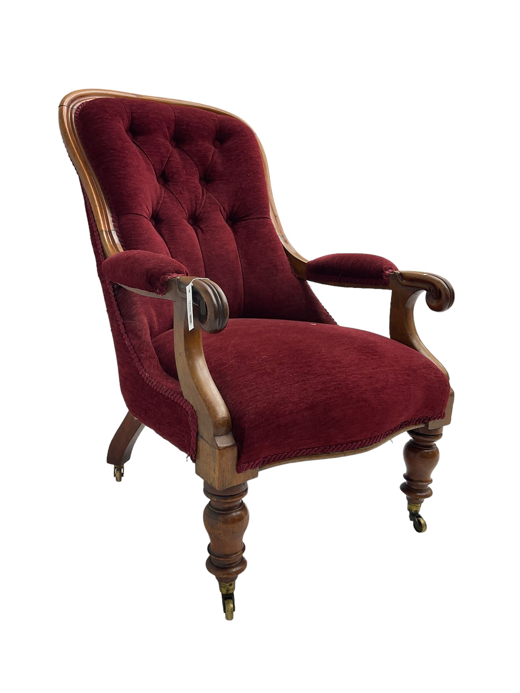 Victorian mahogany framed open armchair, upholstered in buttoned red fabric, the arm terminals carved with scrolls, on turned front supports with brass cups and castors