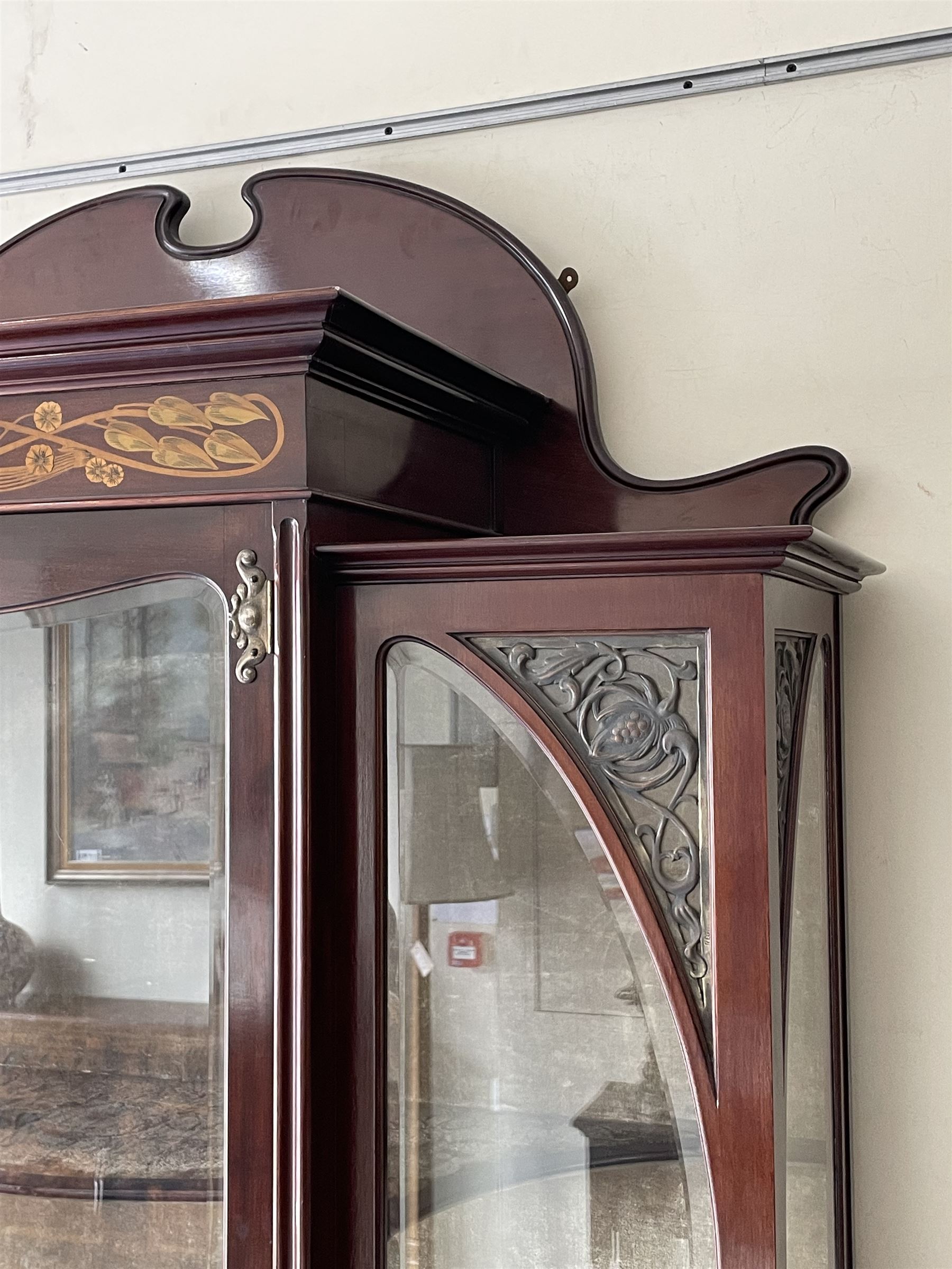 Art Nouveau period inlaid mahogany display cabinet, raised shaped and moulded pediment over projecting moulded cornices, the upper frieze inlaid with trailing branches and tulips, single astragal and bevel glazed door enclosed velvet lined interior fitted with shelves, flanked by curved glass panes decorated with pierced metal foliate spandrels, lower flanking shelves with mirror backs enclosed within shaped and pierced rails inlaid with flower heads, on tapering octagonal supports terminating to compressed pad feet, the lock stamped 'Salmon Bros, London' 