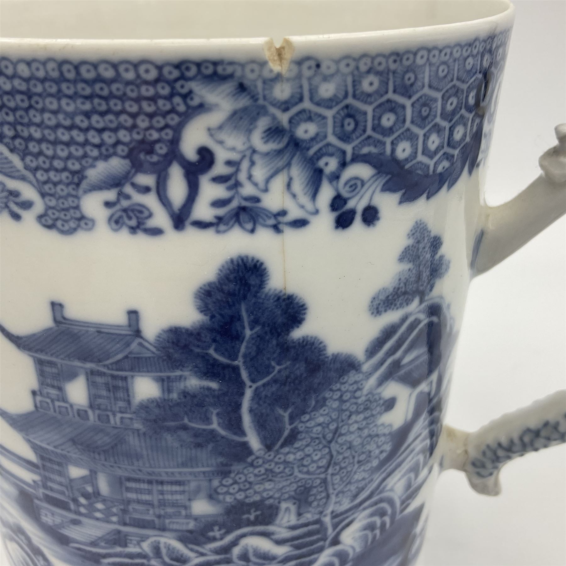 Two large 18th century Chinese blue and white tankards, each of cylindrical form, the first example with foliate mounted strap handle, the body painted with a shaped panel depicting a riverside landscape with pagodas and pine trees, against a textured ground decorated with floral sprays, H15cm D12cm, the second with serpent handle, the body painted with a similar riverside landscape, H14cm D12cm