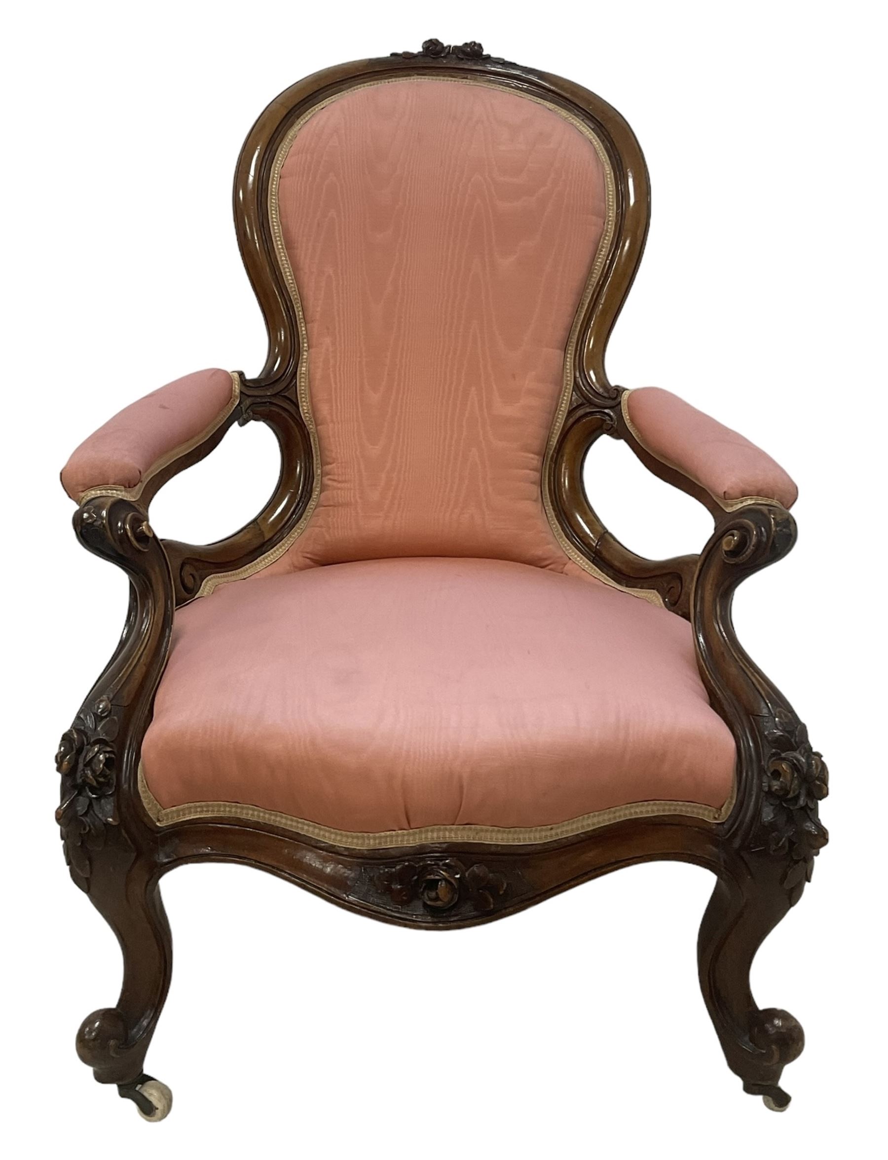 Victorian mahogany framed open armchair, spoon-back and sprung seat upholstered in pink fabric, scrolled arm terminals over serpentine fronted seat decorated with moulded carved floral motifs, cabriole supports with ceramic castors