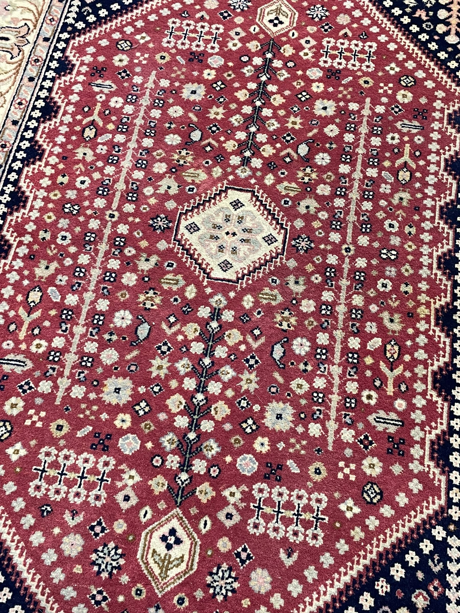 Persian design crimson ground rug, the field decorated with tree of life and floral motifs, enclosed by indigo spandrels with stylised flower head decoration, repeating scrolling border 