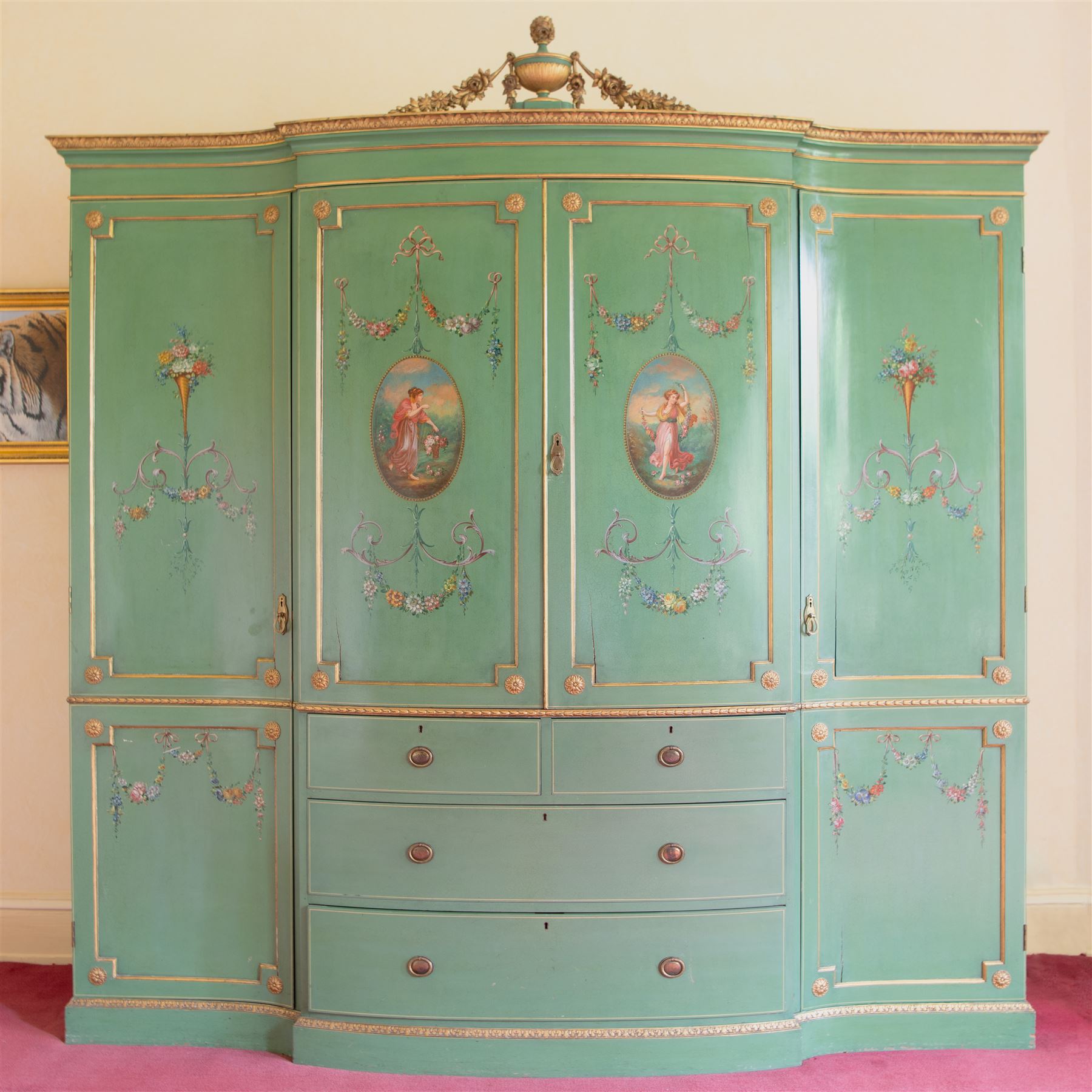 Edwardian Adam Revival break-bowfront quadruple green-painted and parcel gilt wardrobe, the projecting acanthus leaf moulded cornice surmounted by urn with trailing flower heads, two central panelled doors enclosing linen slides, decorated with oval painted panels depicting maidens with flowers and scrolled acanthus leaves with floral festoons, two short and two long drawers below, flanked by full-height panelled doors enclosing hanging rails and hooks, foliate moulded plinth base