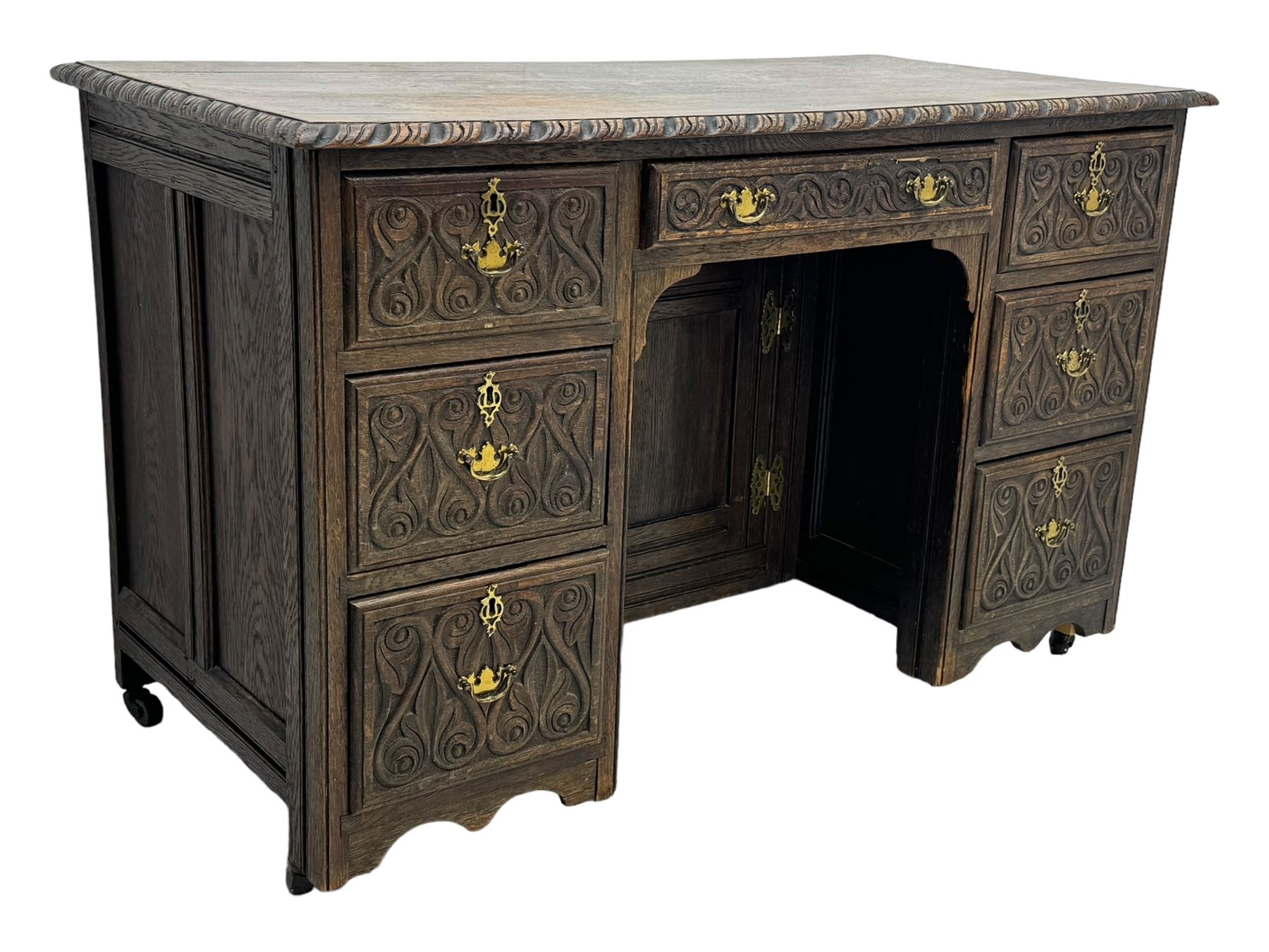 Victorian carved oak kneehole desk, the rectangular top with carved edge, above central drawer with carved front and brass handles, central kneehole with cupboard door, flanked by two banks of three graduating drawers each with similarly carved decoration, on shaped plinth base with castors