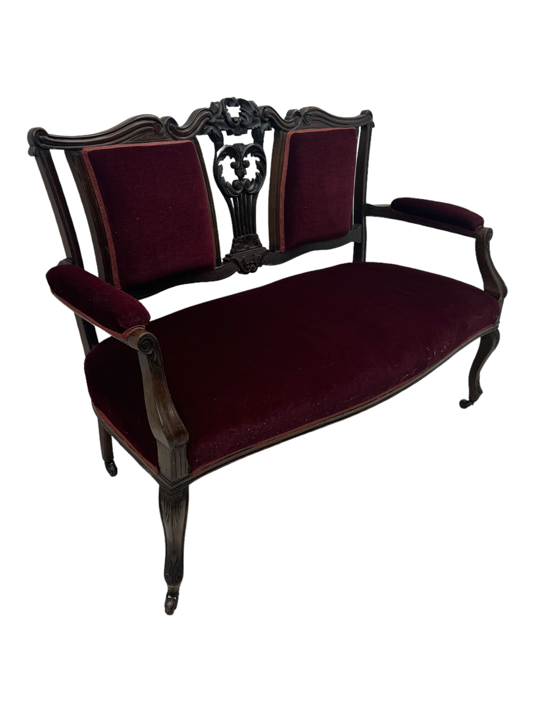 Late Victorian dark oak framed sofa, the backrests, seat and padded arms upholstered in deep red fabric, carved crest rail with scroll and foliate motifs supported by carved uprights, on cabriole supports with leaf carvings terminating in castors