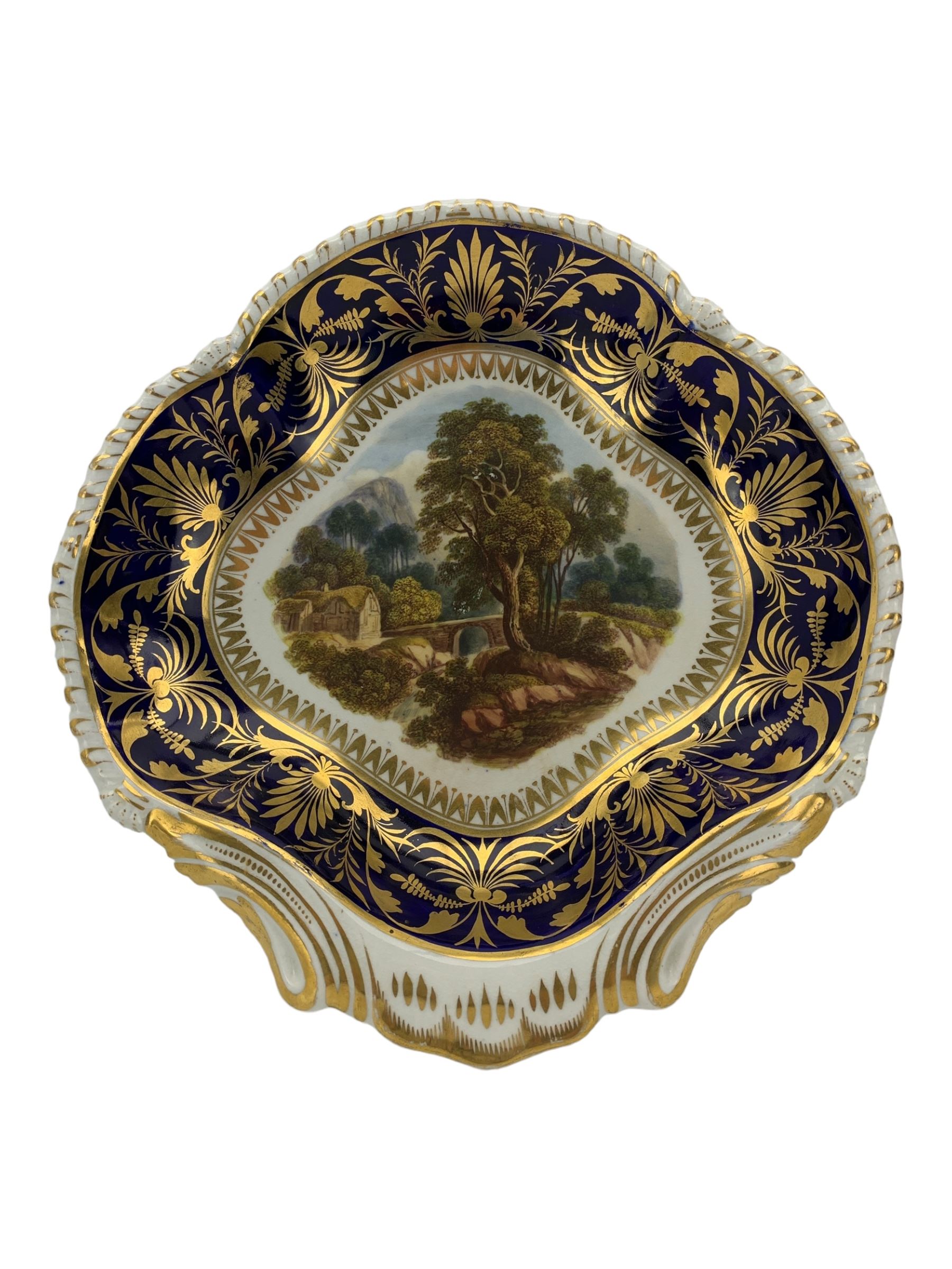 Early 19th century Derby porcelain urn, the reserve painted with a pastoral landscape and castle in the distance, with twin scroll and mask handles, upon a square pedestal base, H23cm, together with a Derby shell-shaped dish, centrally hand painted with a landscape scene titled 'In North Wales', within a cobalt blue and gilt border, L25cm (2)