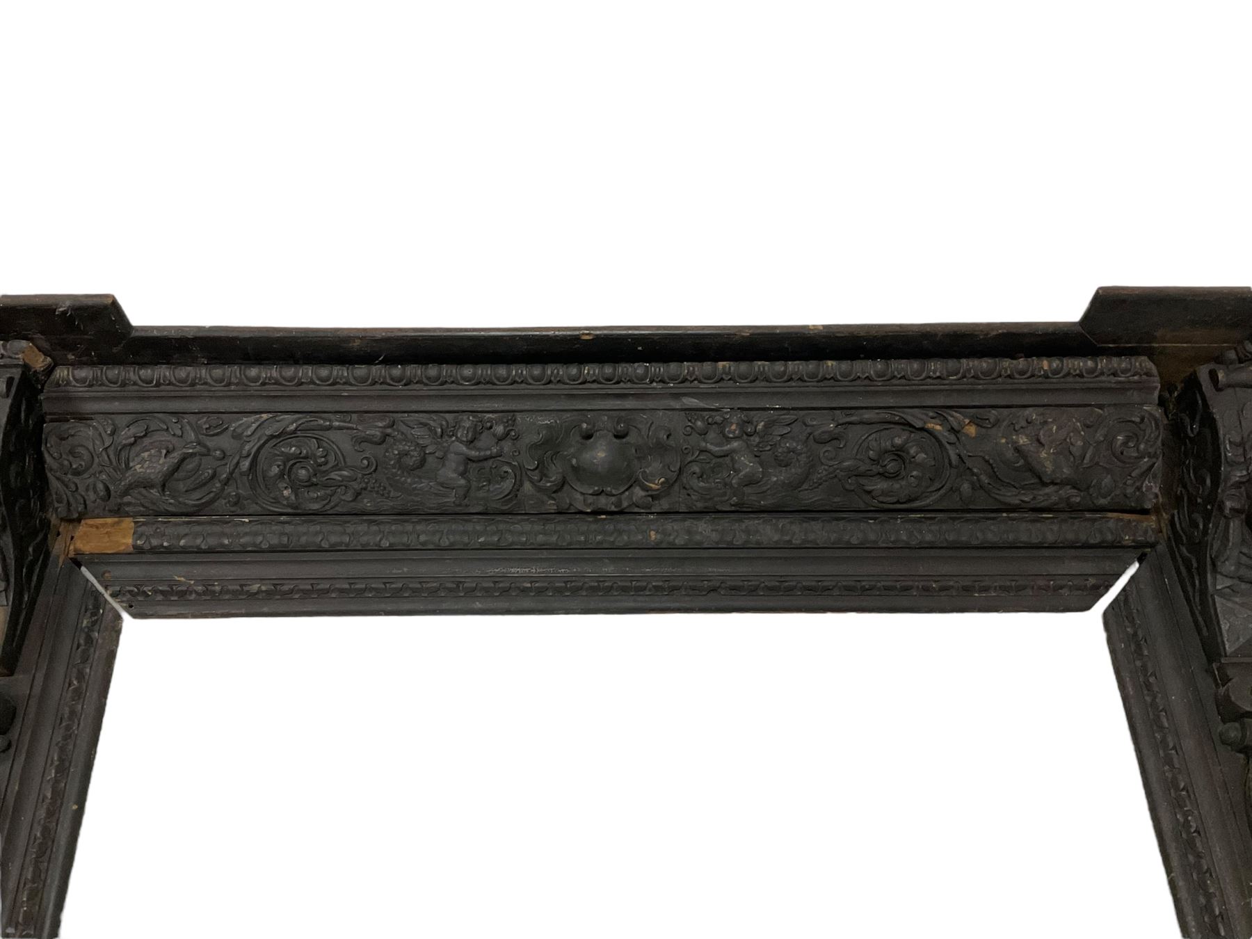 19th century Elizabethan design carved oak fire surround, reverse break-front lintel over egg and dart moulded upper edge, the ovolo-moulded frieze carved with scrolling leafy branches, central cartouche and putti riding dolphins, the uprights carved with female masks over carved busts, square tapering pilasters carved with ribbon tie over trailing fruit, on stepped moulded base 