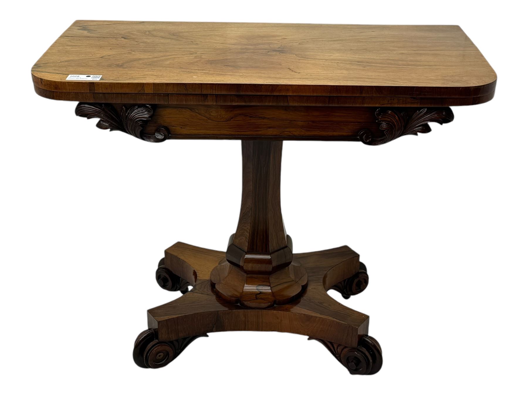 Early Victorian rosewood card table, rectangular foldover swivel top with rounded corners, red baize-lined surface, frieze adorned with carved foliate motifs, supported by a central octagonal waisted column on shaped quatrefoil base, terminating in scrolled feet with concealed metal castors