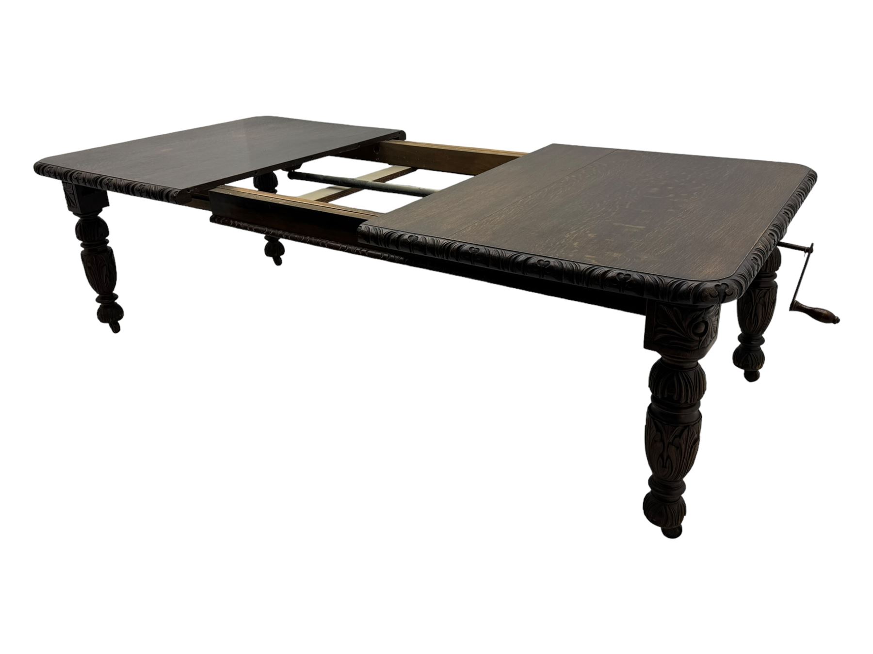 19th century heavily carved oak extending dining table, rectangular top with rounded corners and carved gadrooned edge, extending via winding mechanism with two additional leaves, raised on acanthus leaf-carved baluster supports terminating in ceramic castors