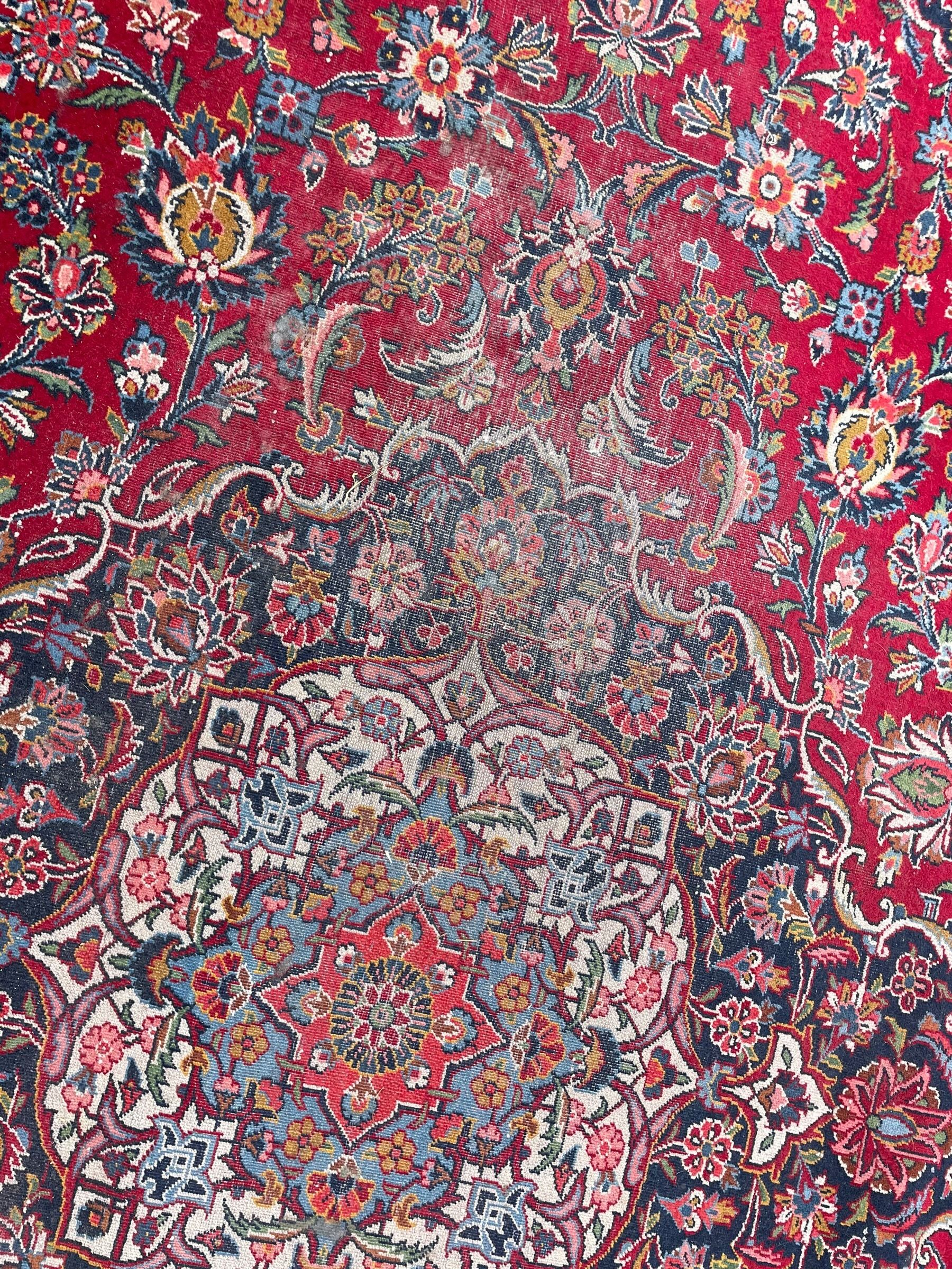 Large Persian Kashan crimson ground carpet, central floral medallion surrounded by swirling leafy branches and palmettes, enclose by floral pattern spandrels, the indigo border with overall scrolling design decorated with palmettes, within guard stripes 